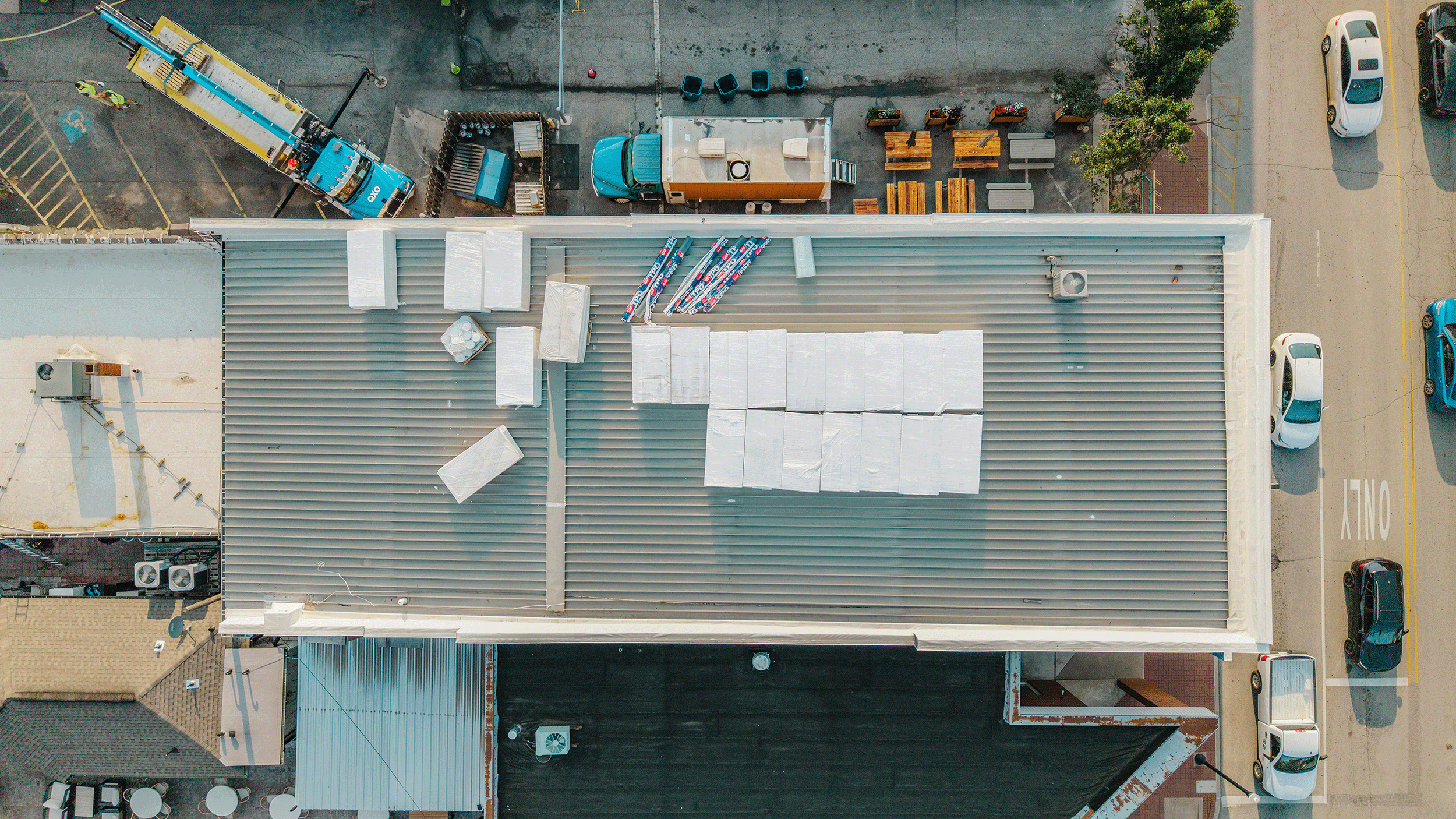 Aerial view of a rooftop with white insulation material, an air conditioning unit, and several piles of insulation rolls. Below the rooftop, a parking lot with parked cars and a small outdoor seating area with tables and benches are visible.