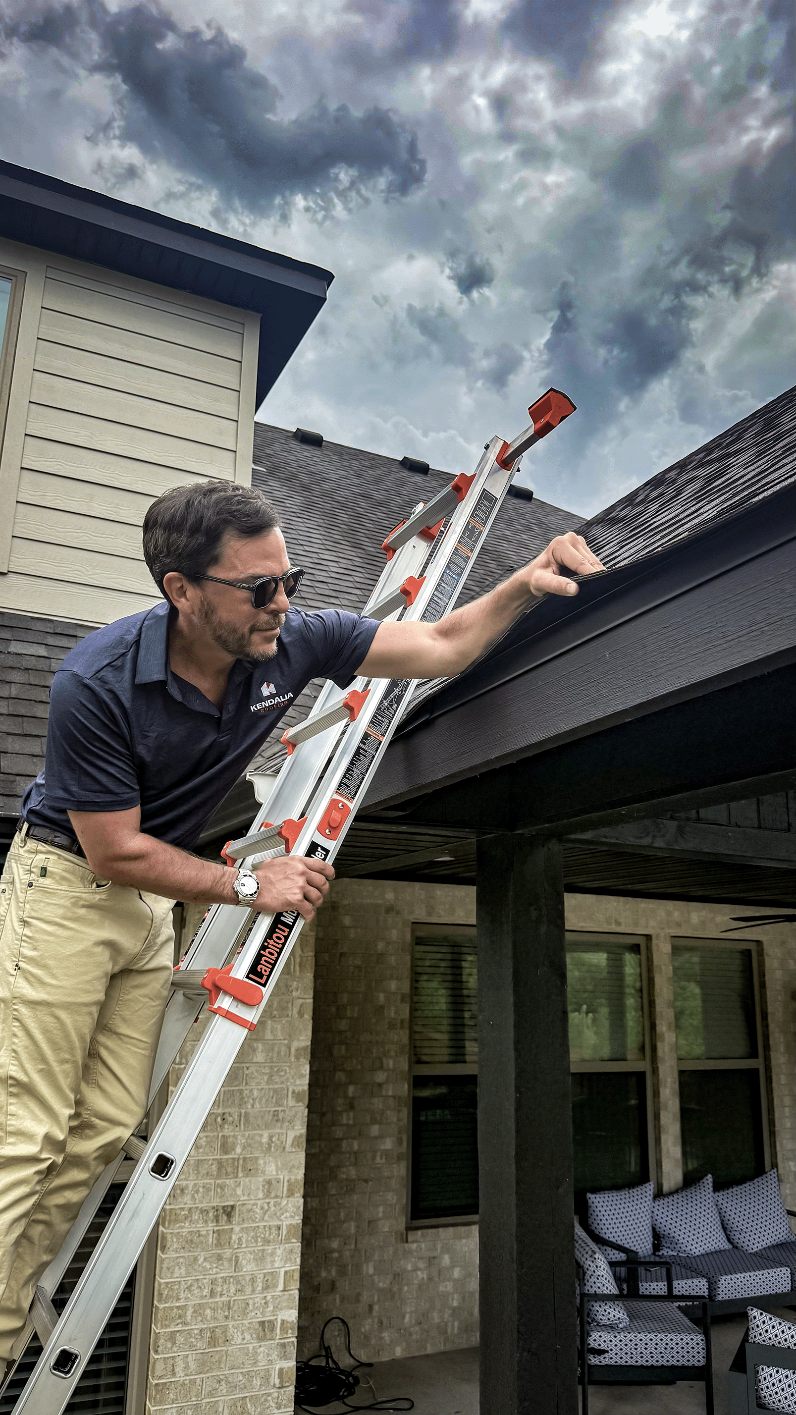 A man in sunglasses and a navy blue shirt is inspecting a dark-colored roof while standing on an extension ladder outside a house. The house has brick walls and a screened porch with outdoor furniture underneath. The sky is cloudy, indicating an overcast day.