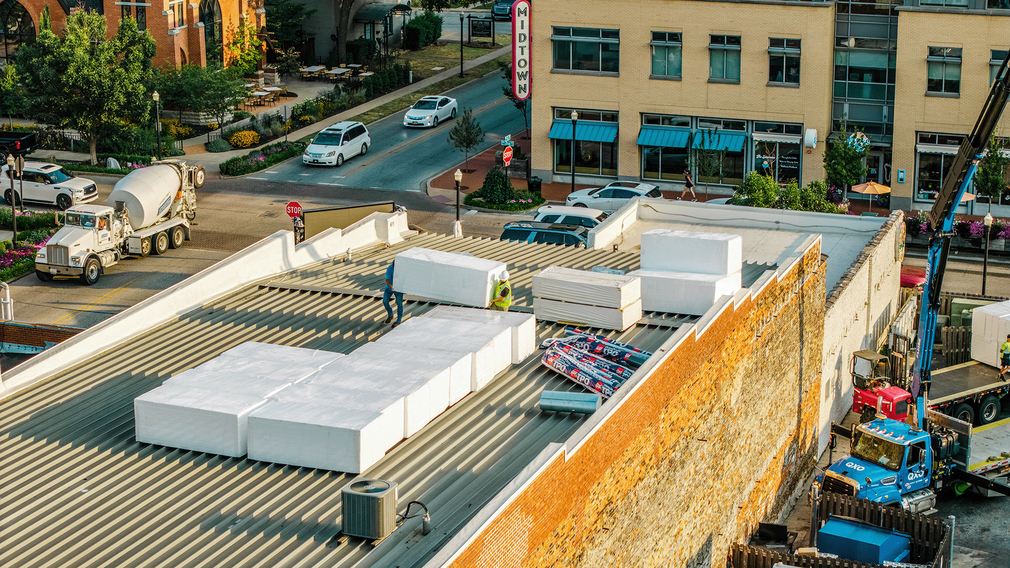 Workers installing foam insulation on the roof of a building with construction equipment nearby, and a busy street with cars and storefronts in the background.