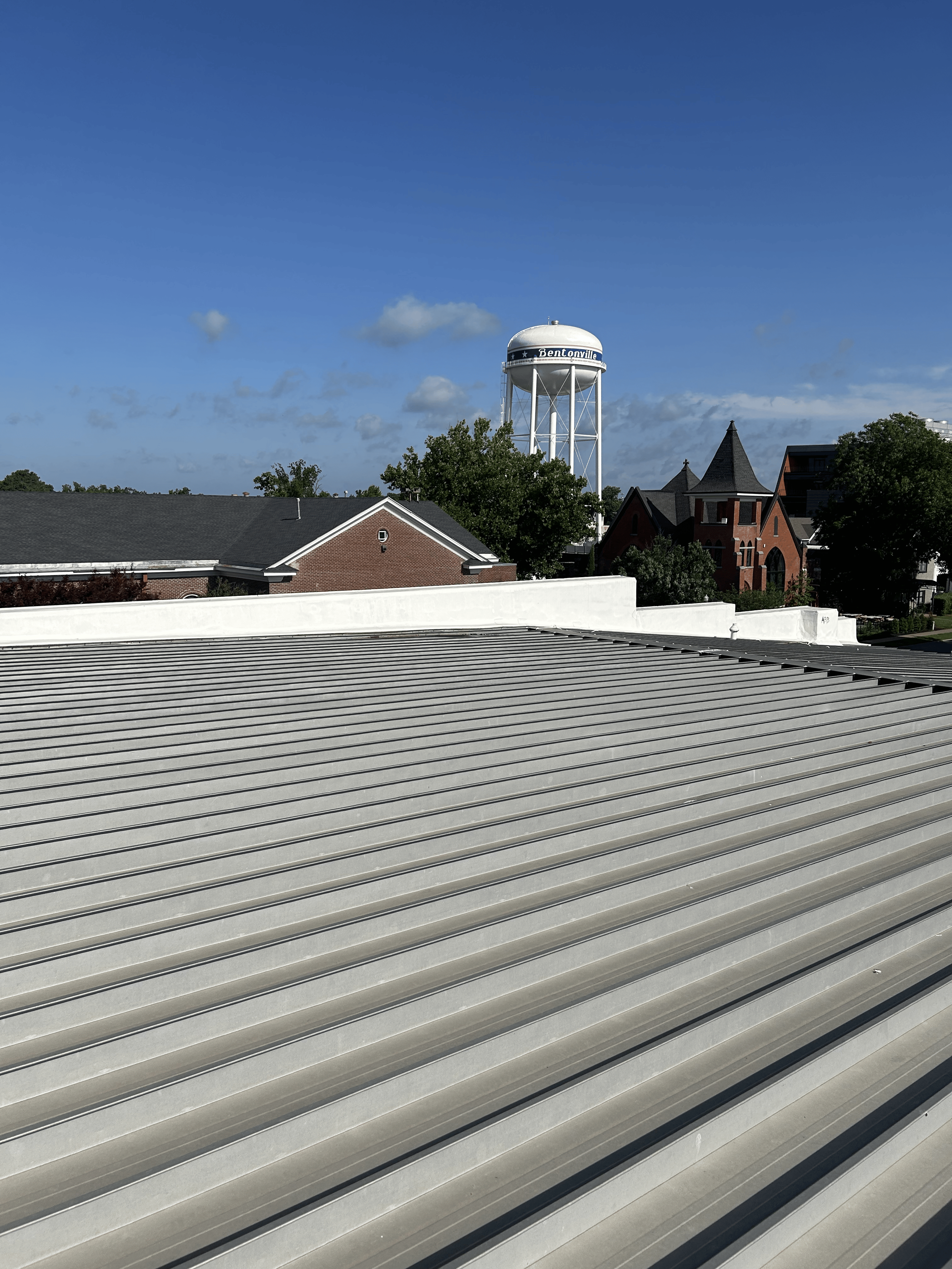 View of a metal rooftop in the foreground with parallel ridges, trees, houses, and a water tower with the label "Bentonville" under a blue sky with a few clouds.