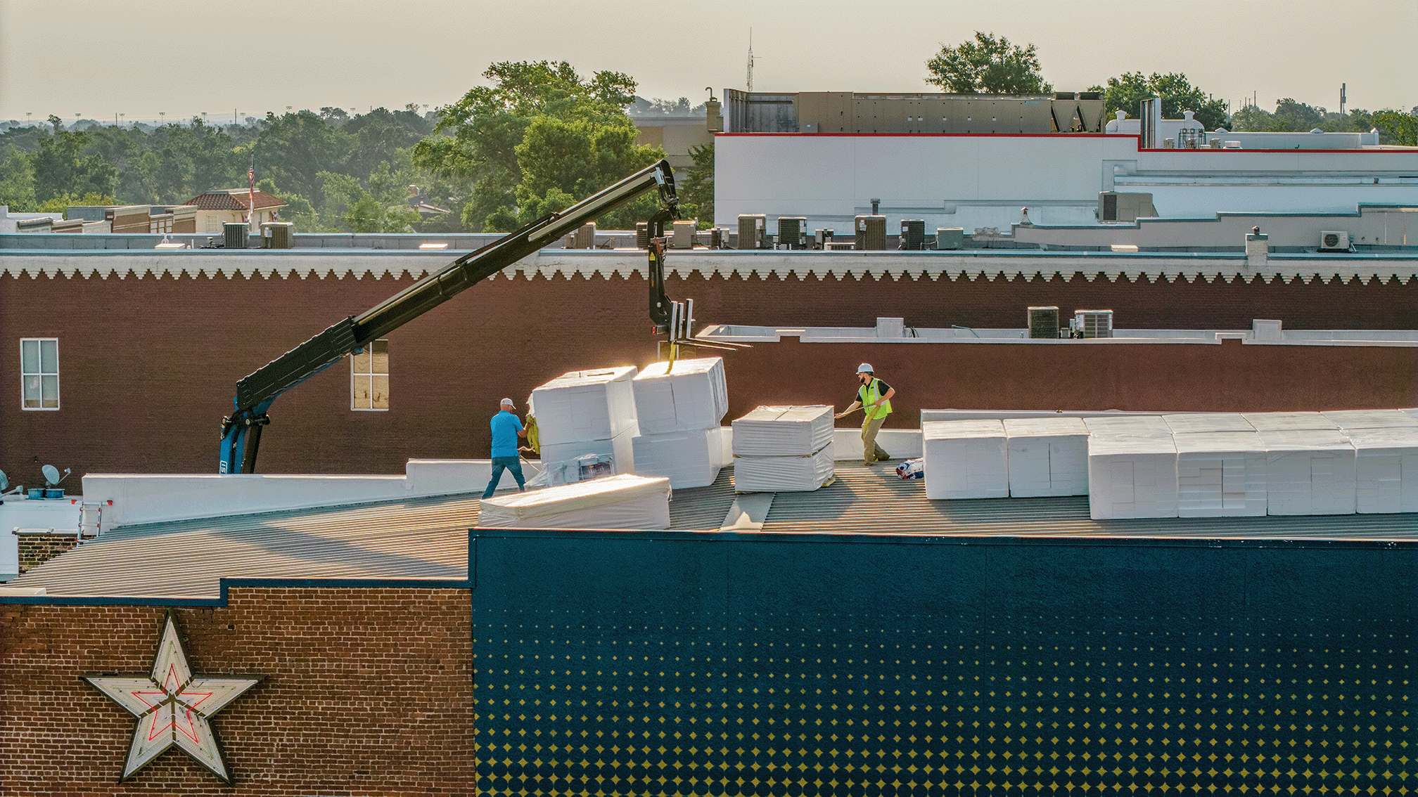 Two workers on a rooftop installing large blocks of white insulation or foam boards, with a crane assisting, in an urban area with trees and buildings in the background.