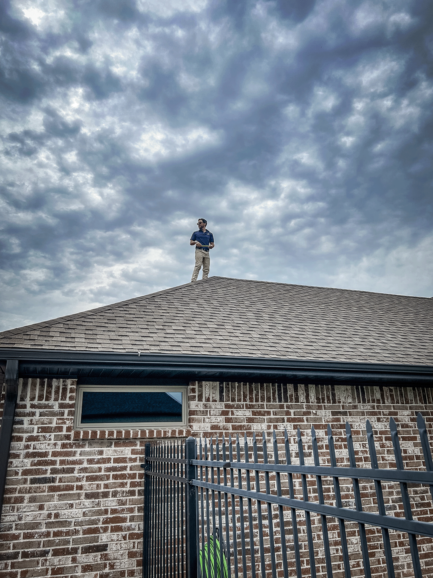 A man standing on a house's roof, overlooking cloudy sky, wearing a navy polo shirt and beige pants, with a black fence in front of a brick wall and a window below.