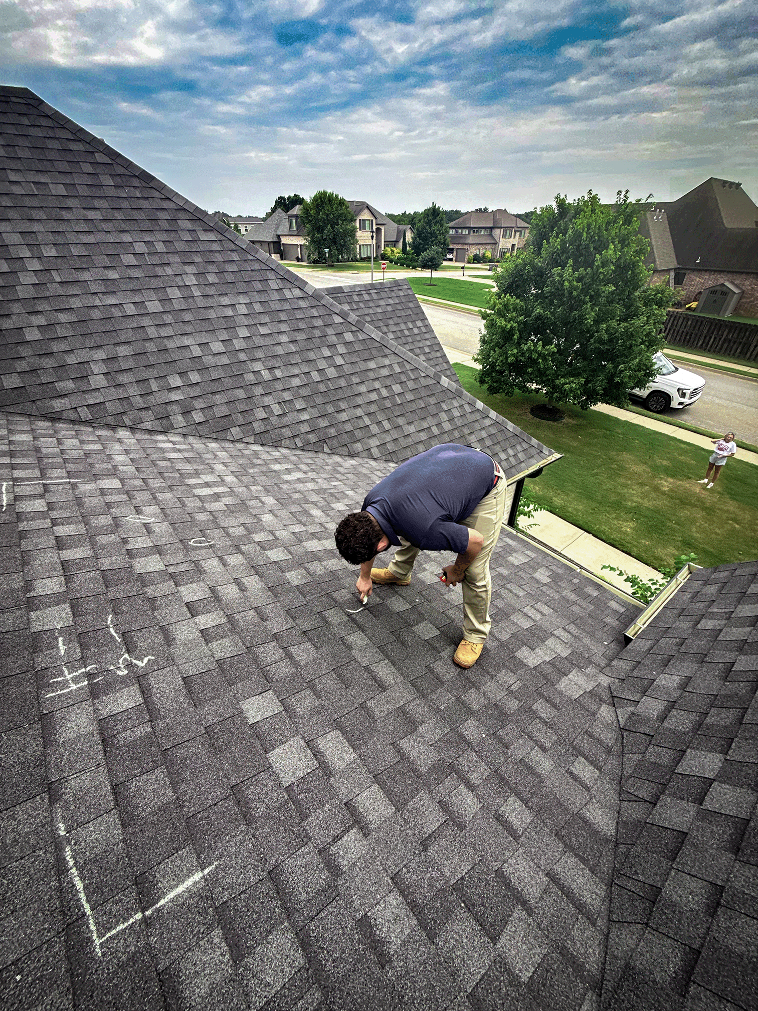 Person installing or repairing shingles on a residential roof, with chalk markings visible on the shingles, in a suburban neighborhood with houses, trees, and a woman standing on the lawn.