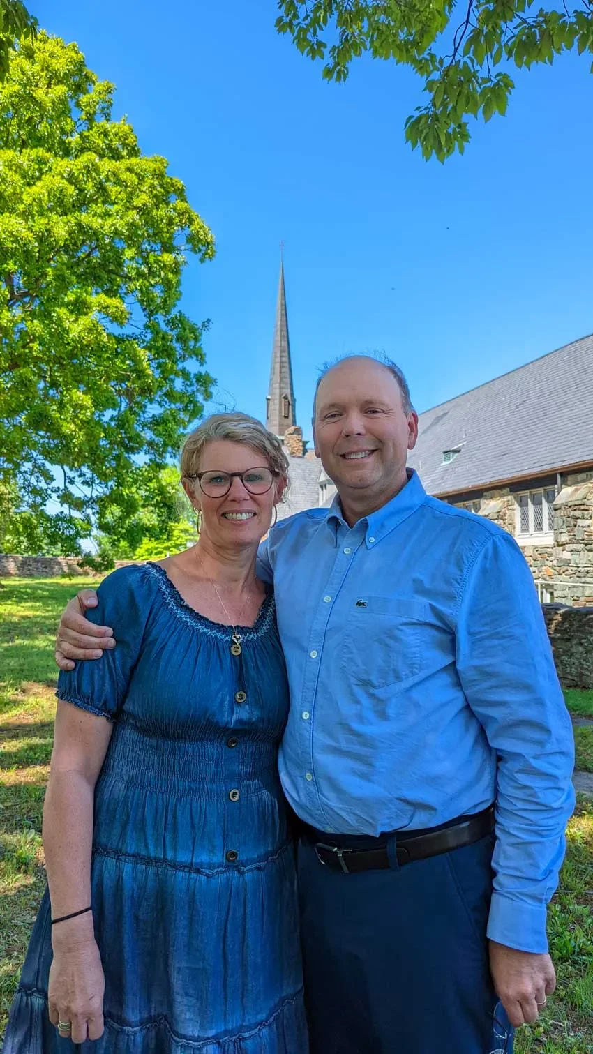 A smiling woman and man standing outdoors with trees and a church steeple in the background.