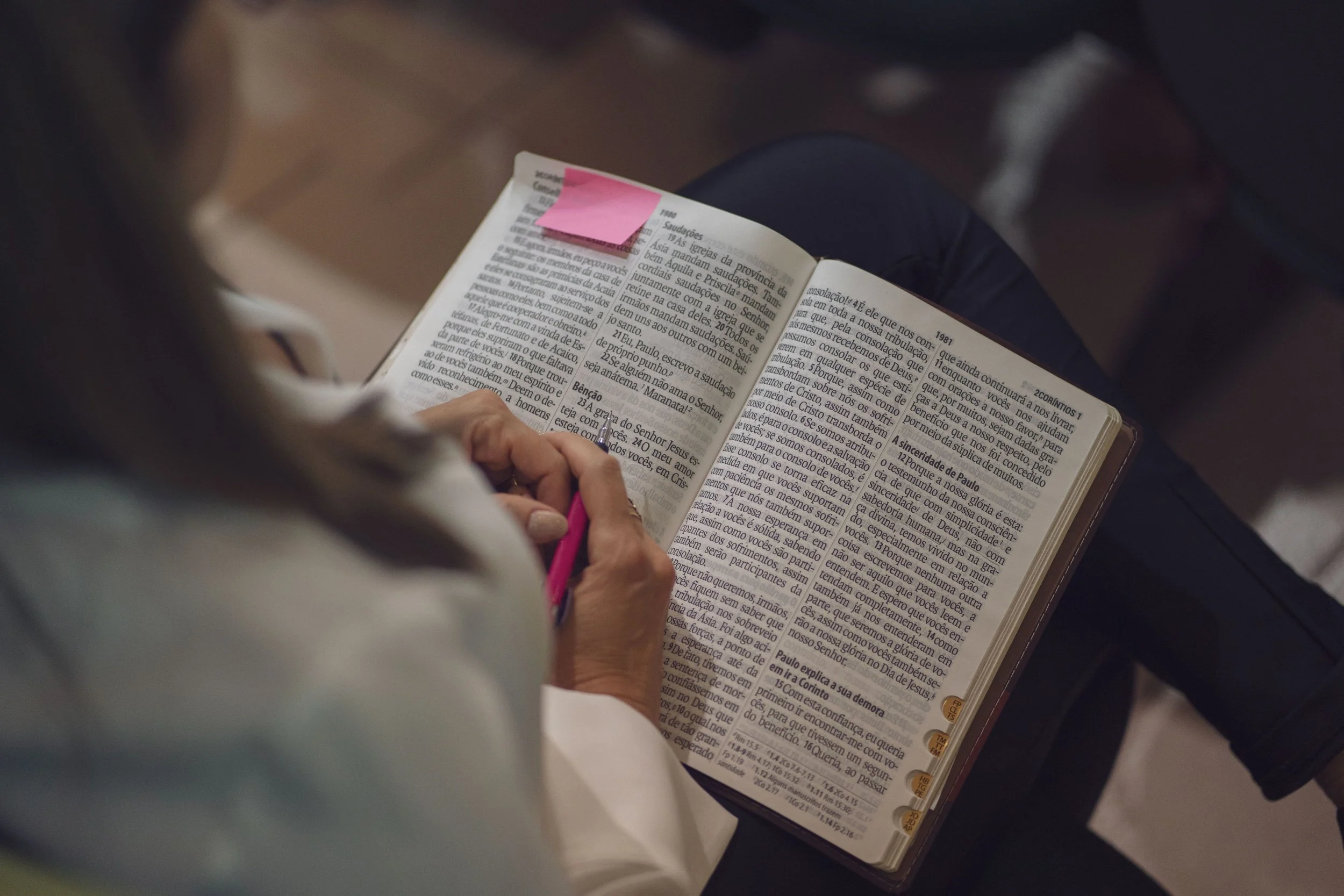 A person holding a Bible and writing with a pink pen, with pink sticky notes attached to the pages.