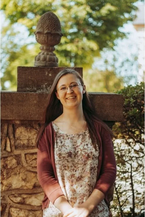 A woman with long brown hair and glasses smiling outdoors in front of a stone and brick structure with a large decorative stone finial on top, surrounded by trees and greenery.