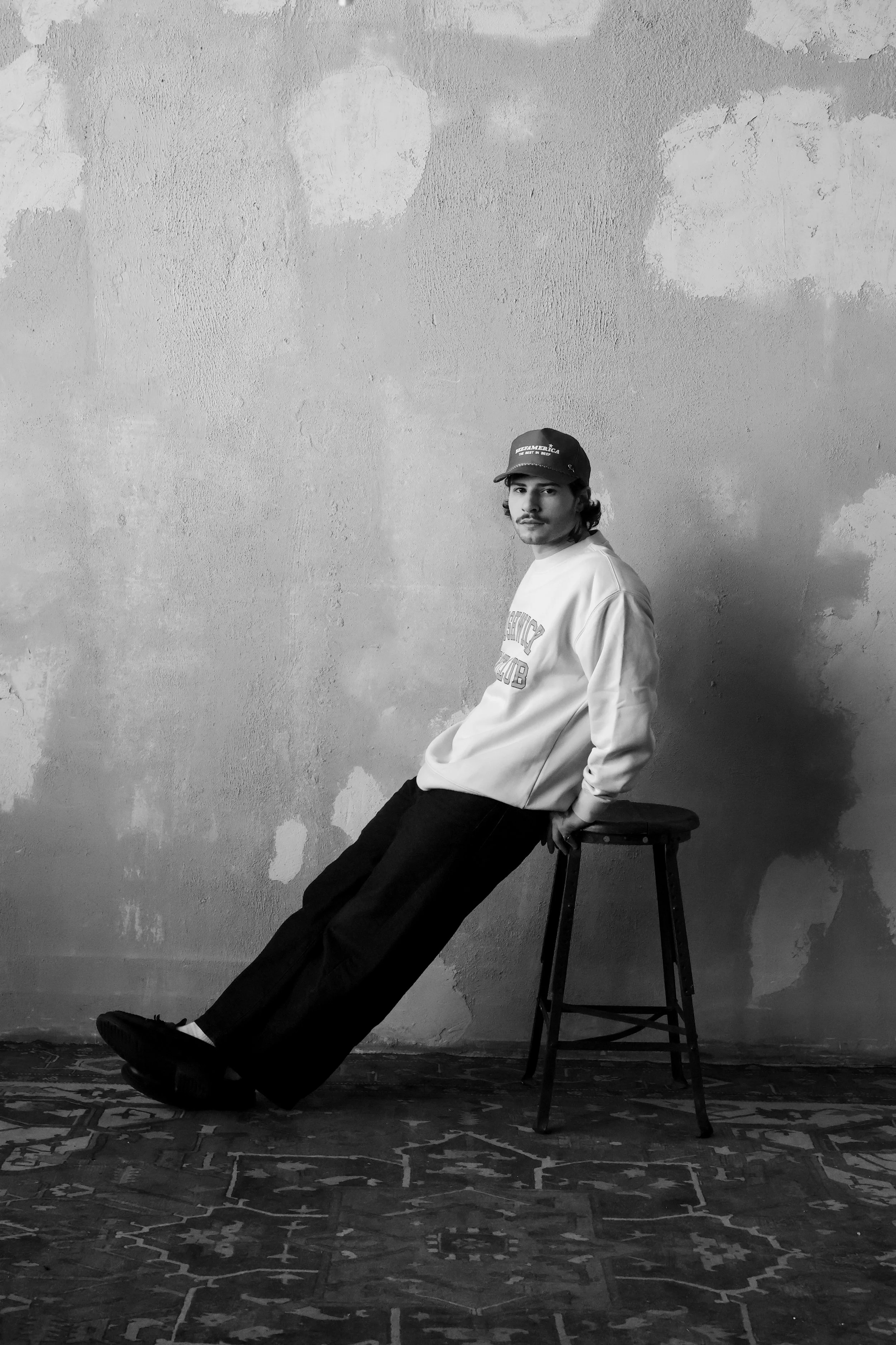 A black-and-white photo of a young man sitting on a stool with his legs extended downward, leaning against a textured wall. He wears a cap, a sweatshirt, and dark pants. The floor has a detailed patterned rug.