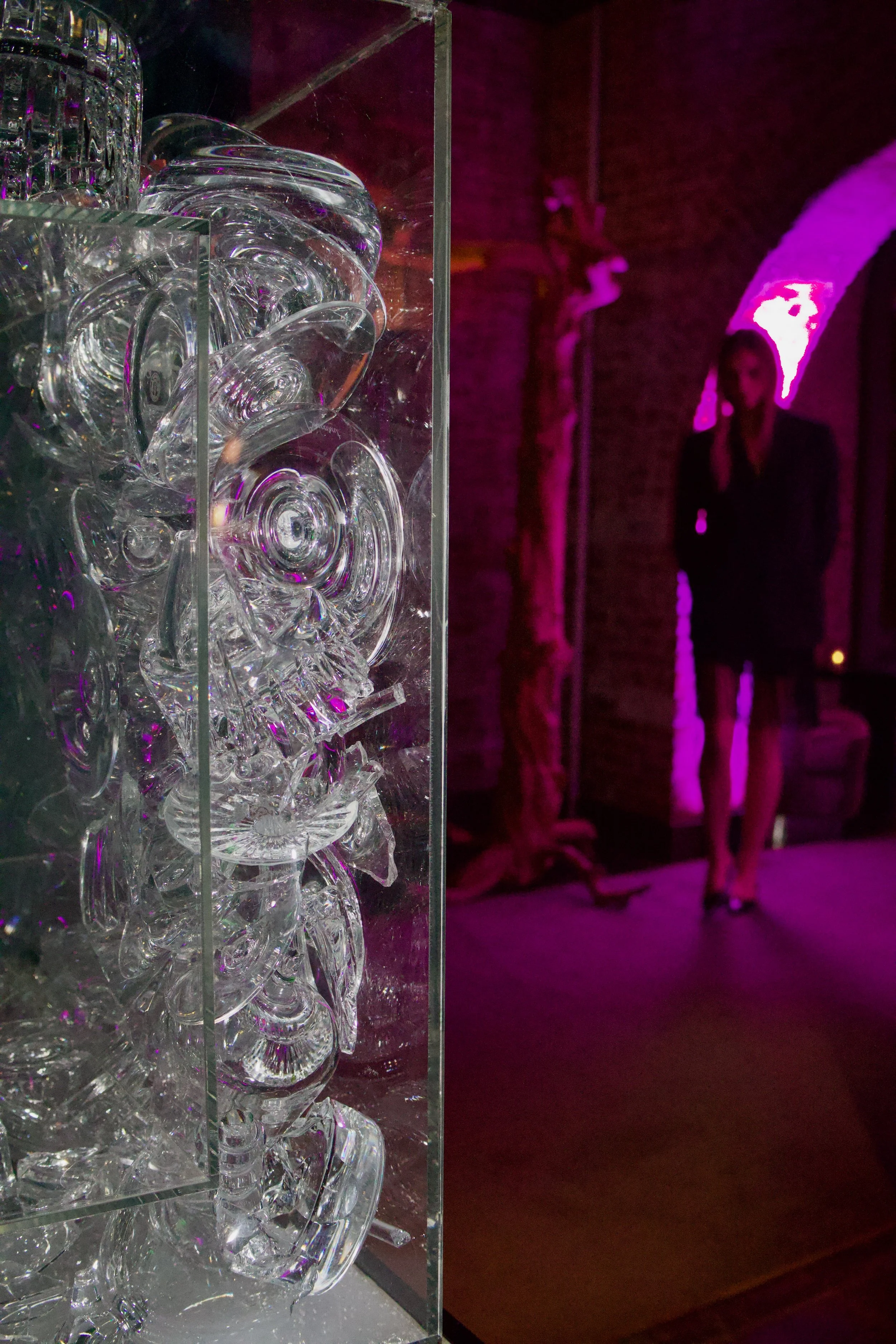 A glass display case filled with clear glass bowls, cups, and decorative glass items, with a pink and purple lit background and a blurred person in a blazer and skirt standing nearby.