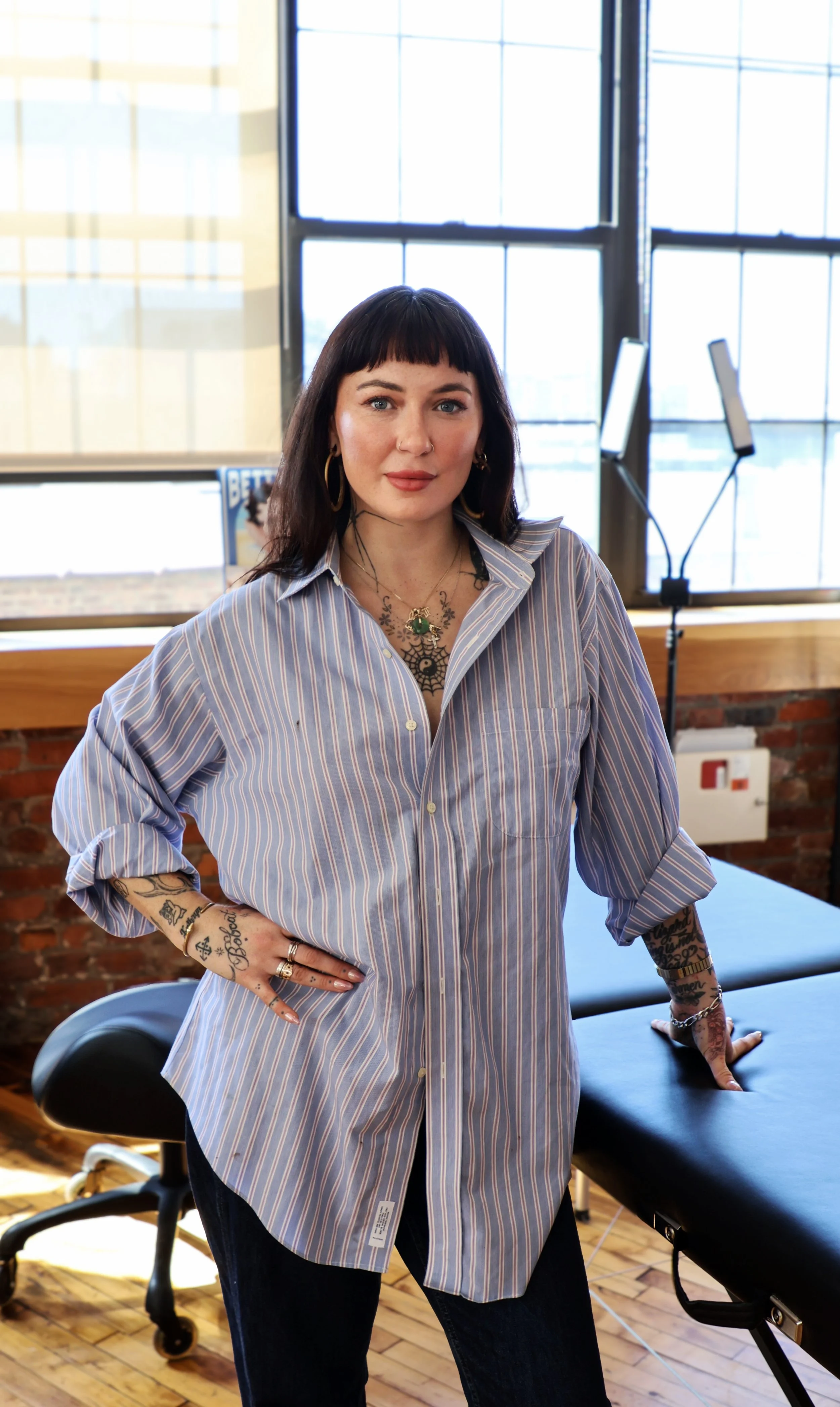 A woman with dark hair and tattoos on her arms and chest, wearing a striped button-up shirt, standing with her hand on her hip in a bright room with large windows, a massage table, and a computer monitor in the background.