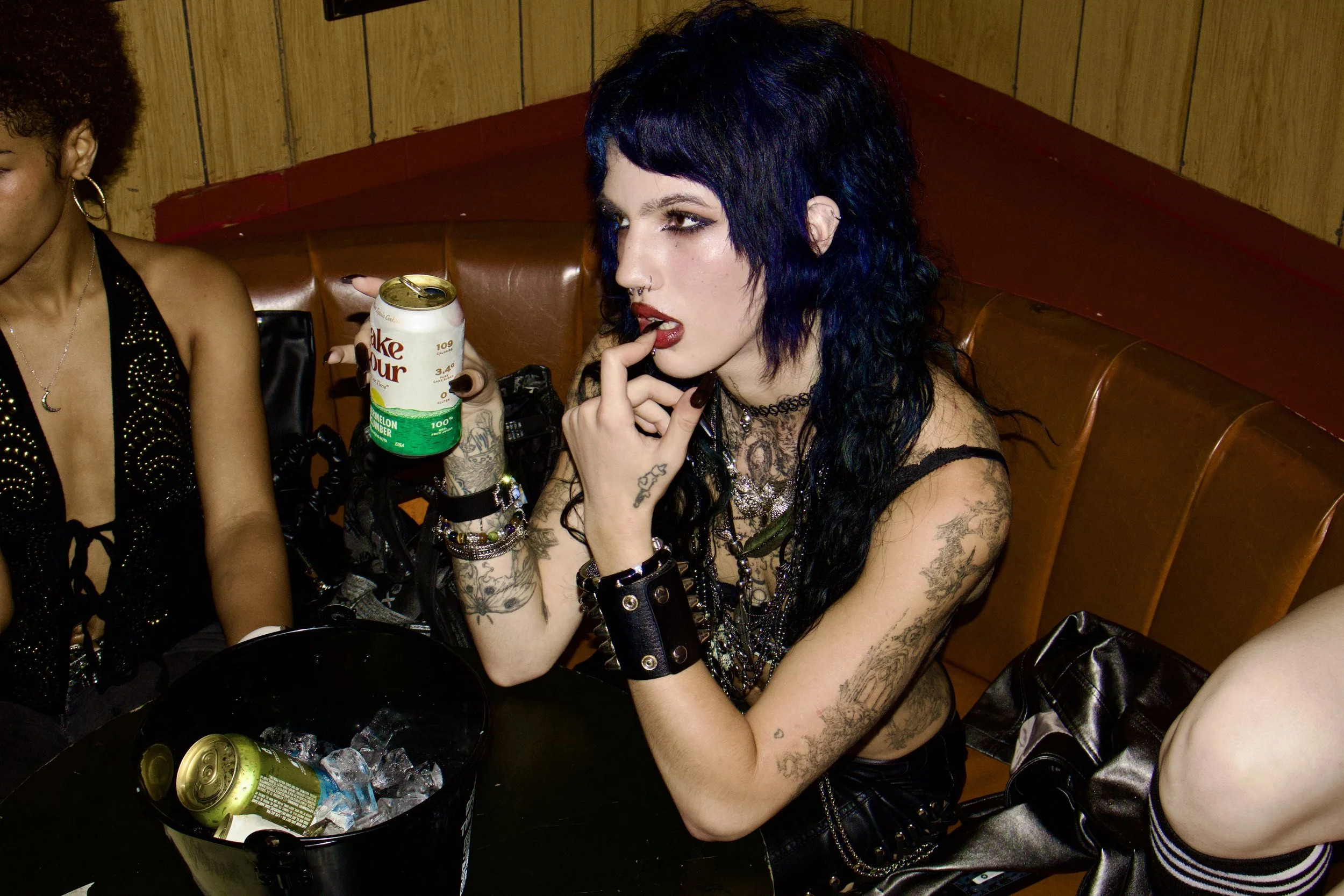 A woman with black hair and tattoos on her arms sitting at a table holding a can of Sprite melon cucumber soda, with a bowl of ice and cans, in a setting with wooden paneled walls.