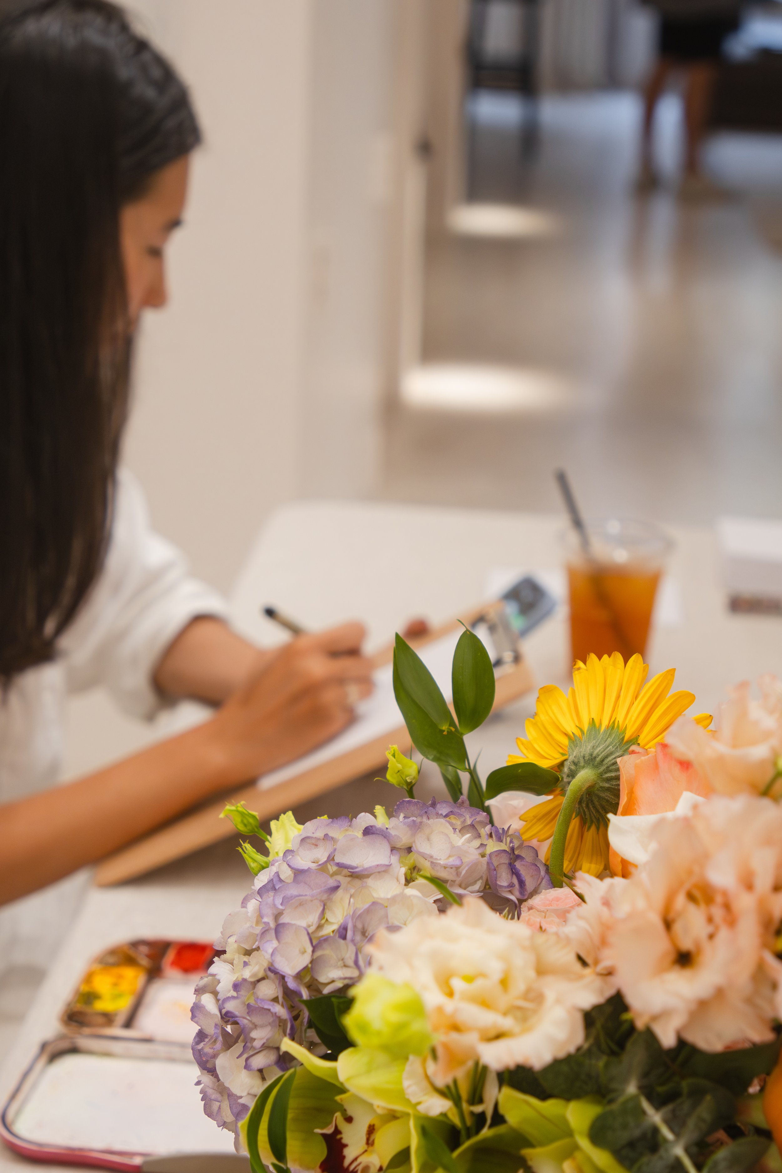 A woman with long dark hair and a black headband, sitting at a table, focused on her phone, with a vase of colorful flowers and a glass of iced tea in front of her in a well-lit room with a wooden floor.