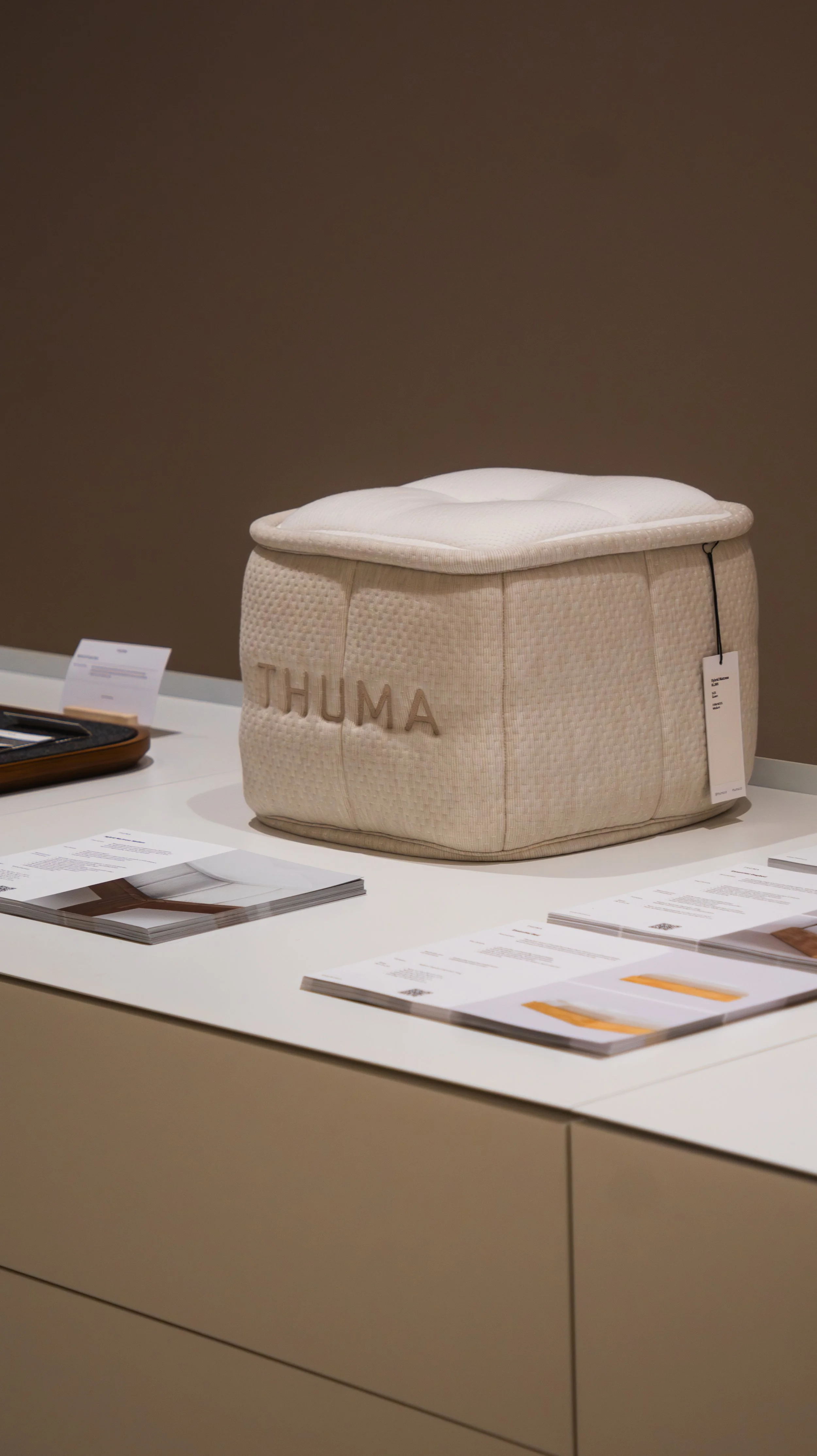 A beige cushioned ottoman with the word 'THUMA' stitched on the side, displayed on a white table with informational brochures and papers around it, against a plain brown wall background.