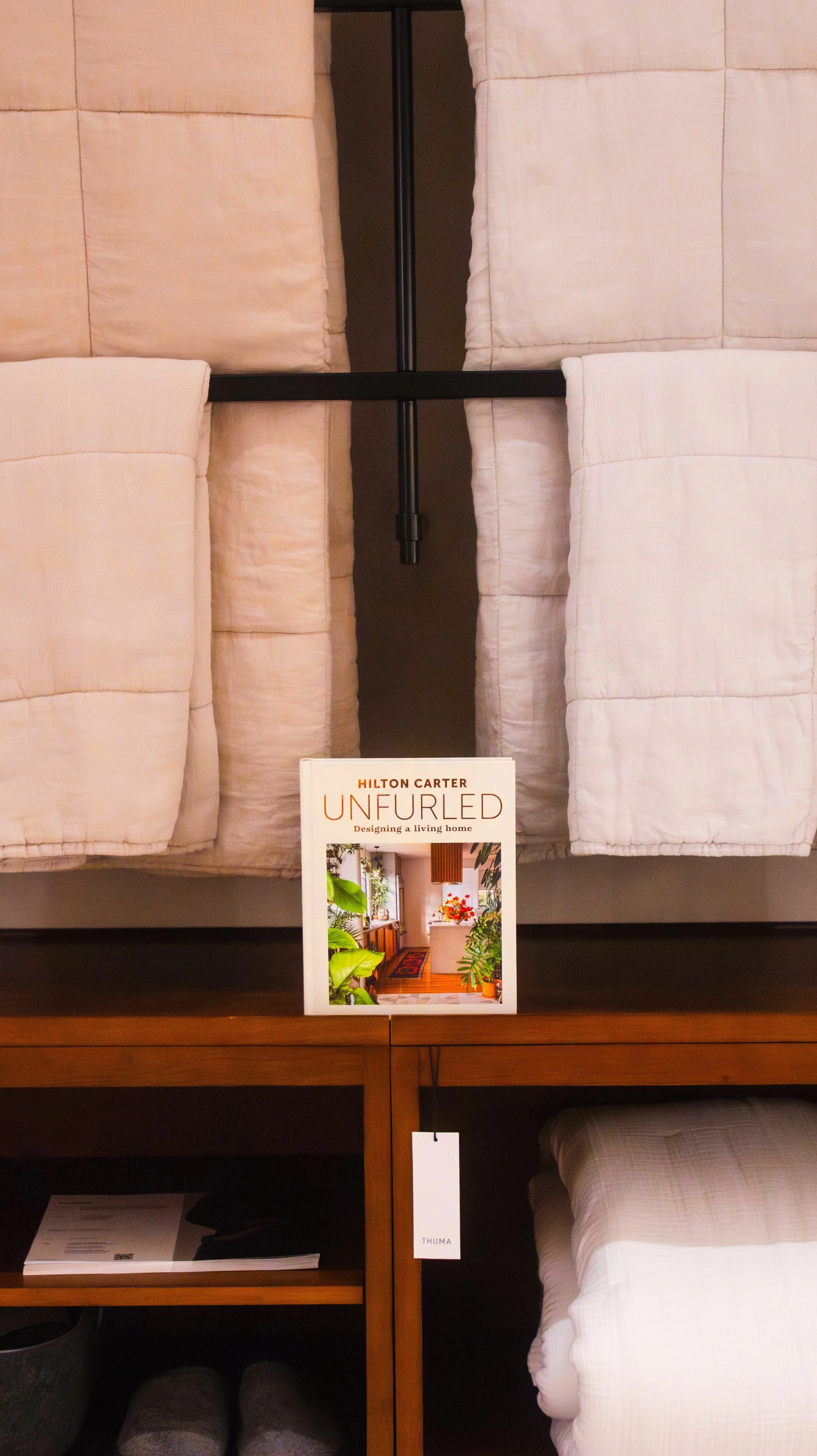 A display of bedroom furniture, including a bed with beige bedding, a wooden side table with a magazine and a tag, and a sign for a book or product titled 'Unfurling: Designing a Living Home' by Hilton Carter. The sign features a photo of a cozy interior with plants and colorful accents.