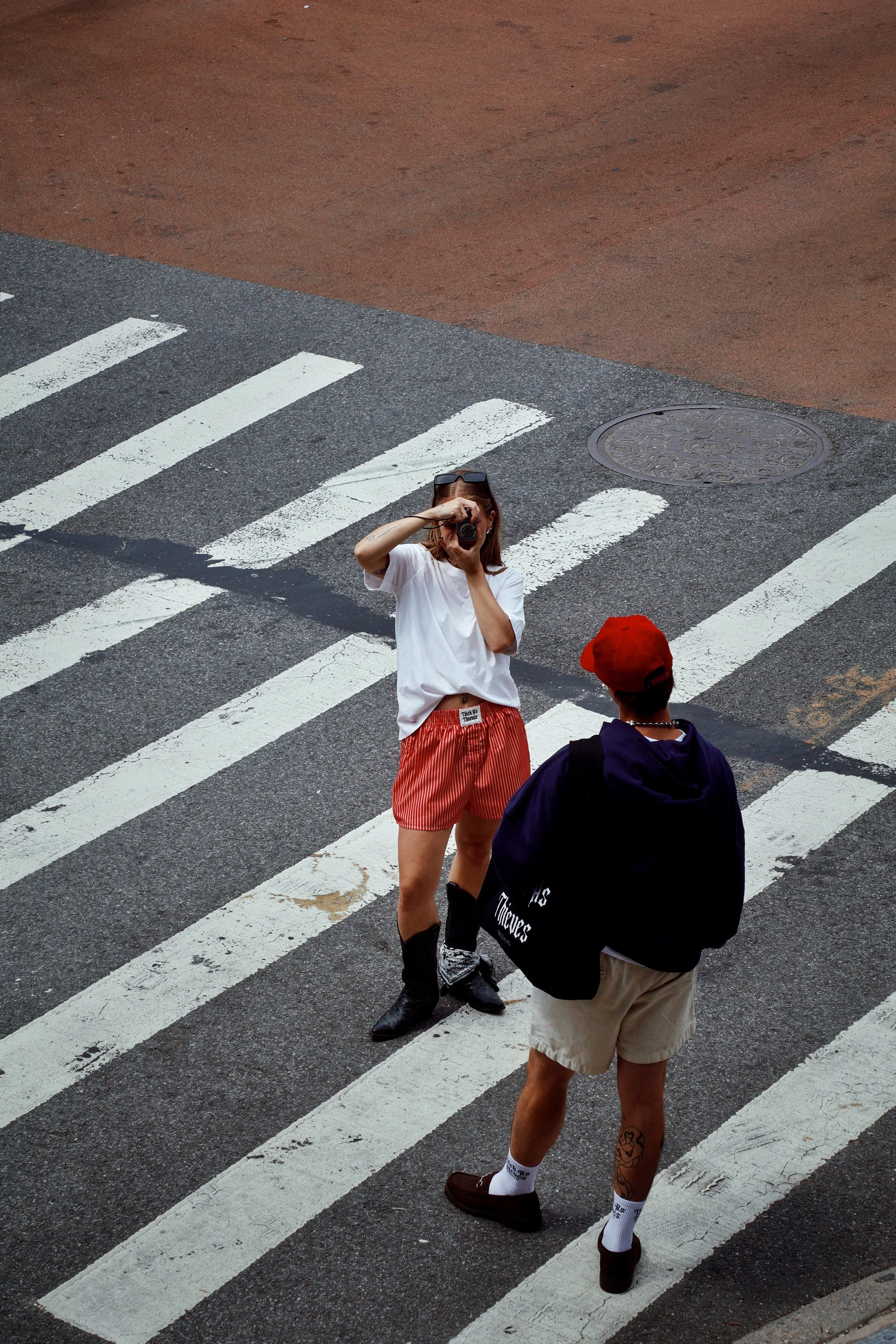 A woman with sunglasses on her head taking a photo with a camera, standing on a striped crosswalk, wearing a white t-shirt and red striped shorts. Another person with a black bag, beige shorts, and a red cap is facing her, with only their back visible.