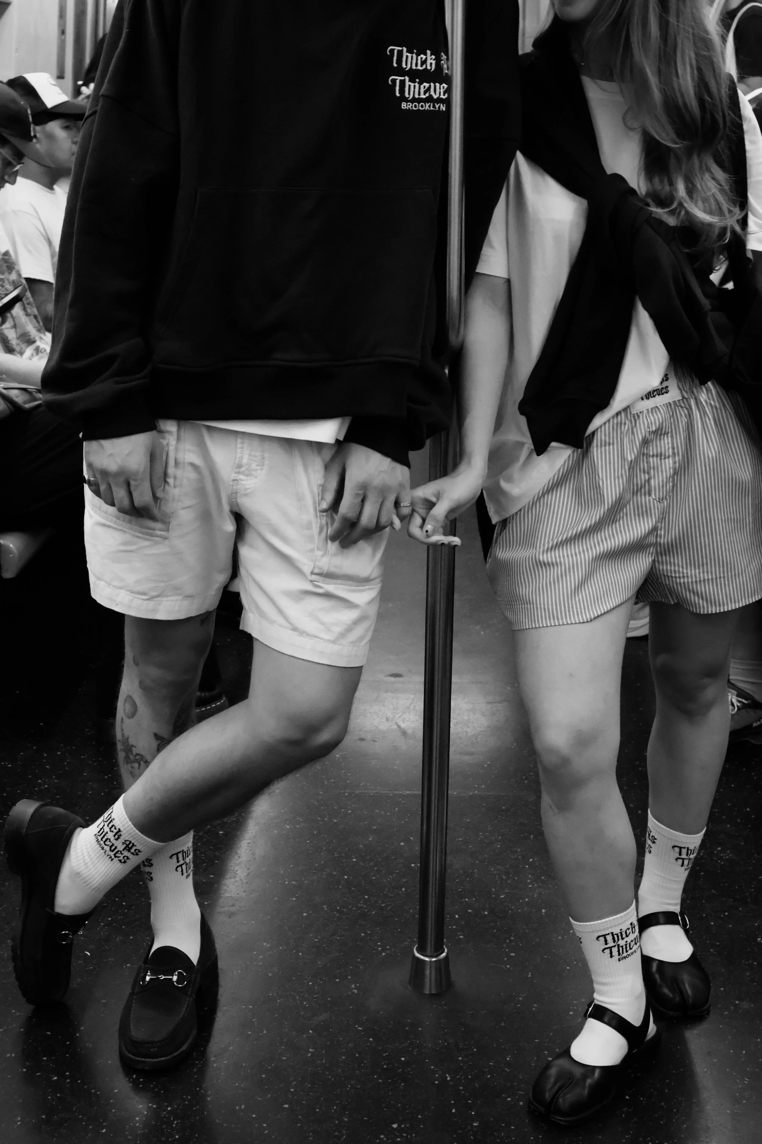 Two people holding hands on a subway, wearing sneakers and matching socks that say "Thick Thief Brooklyn." They have colorful tattoos on their legs and are standing close together.