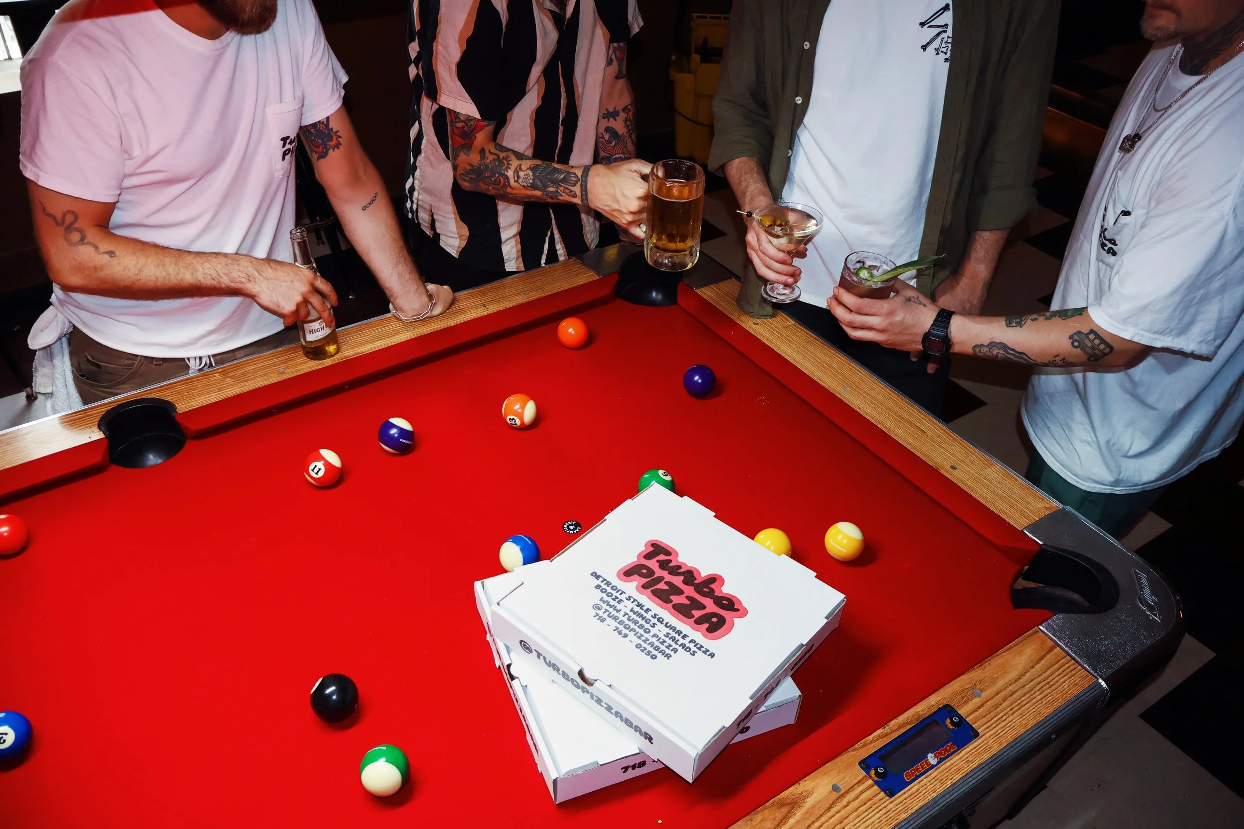 Four men are standing around a red pool table, holding drinks, with pool balls set up for a game. They are dressed casually, some with tattoos, and are engaged in conversation.