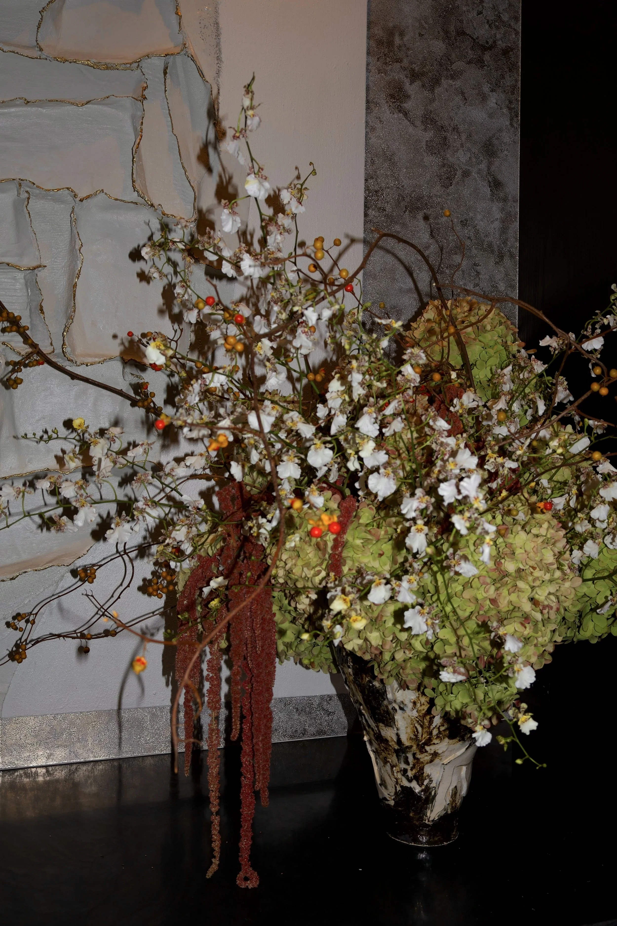 A floral arrangement in a tall, painted ceramic vase featuring white flowers, red and yellow berries, and orange hanging tassels, placed on a dark surface against a textured wall.