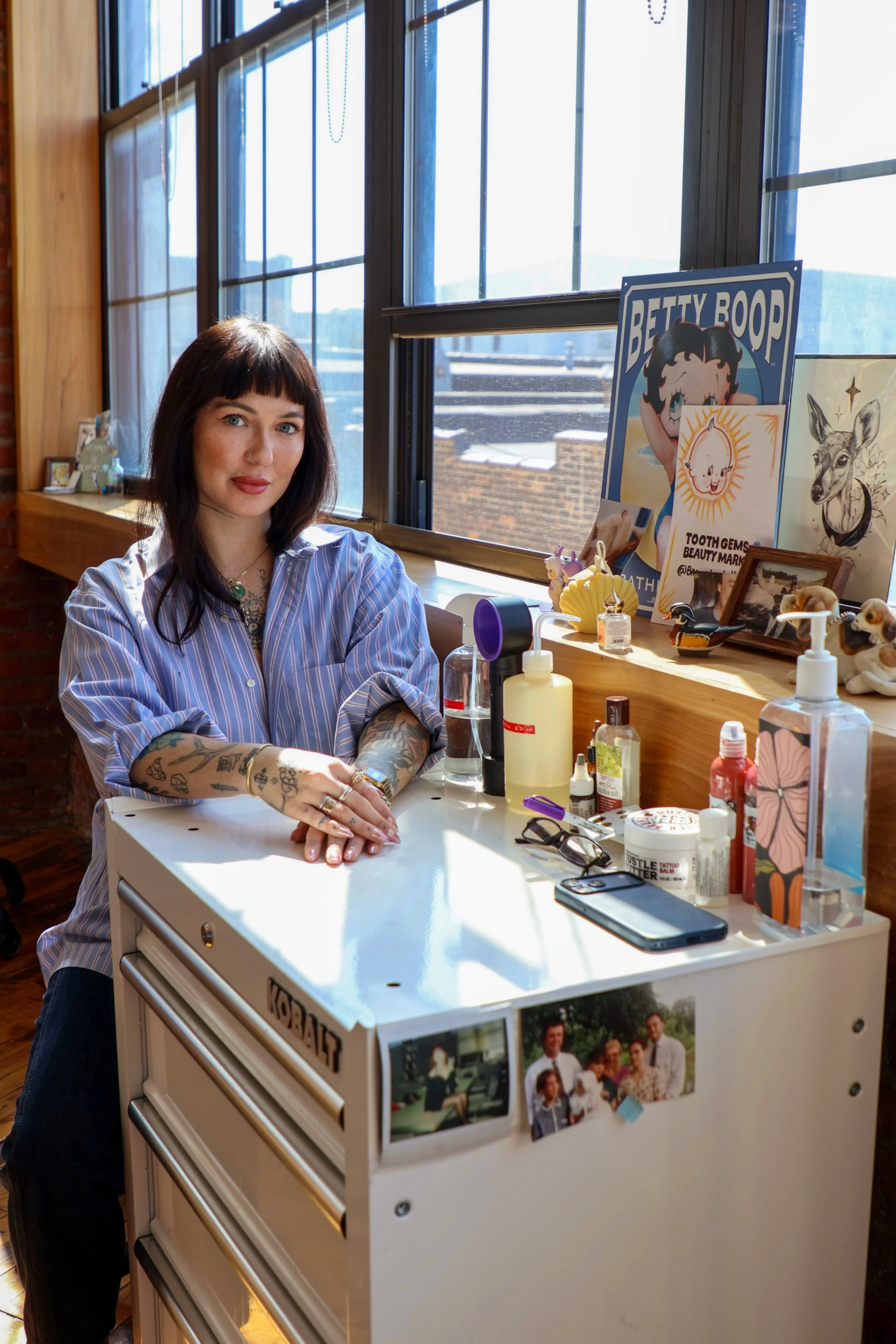 A woman with dark hair, tattoos on her arms, and wearing a striped shirt is sitting at a white vanity table with hair and beauty products. The background shows large industrial windows and decorative posters, including Betty Boop and artwork, with sunlight streaming in.