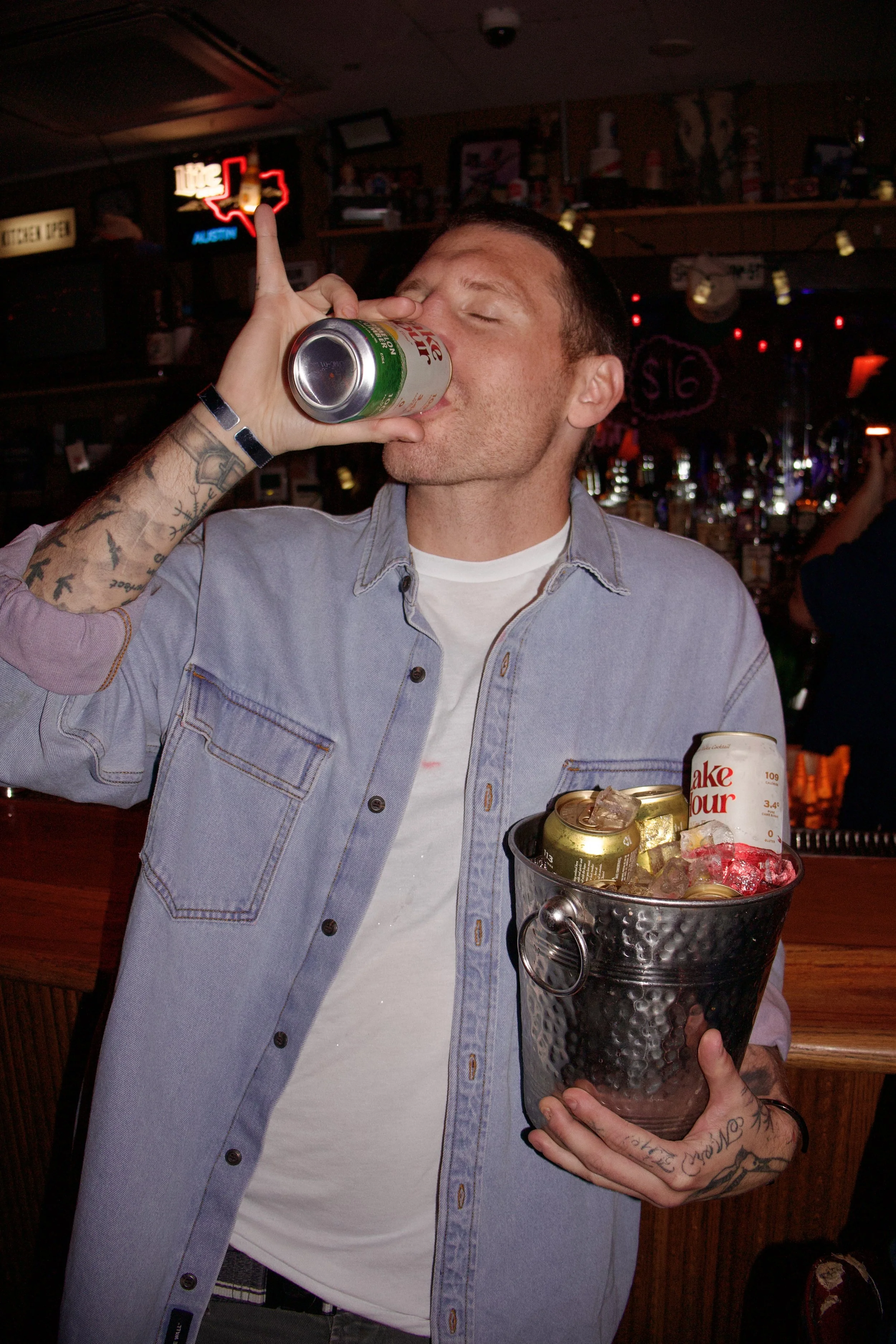 A man with tattoos on his arm holding a bucket with drink cans and ice, while another person drinks from a can in a bar or pub setting.