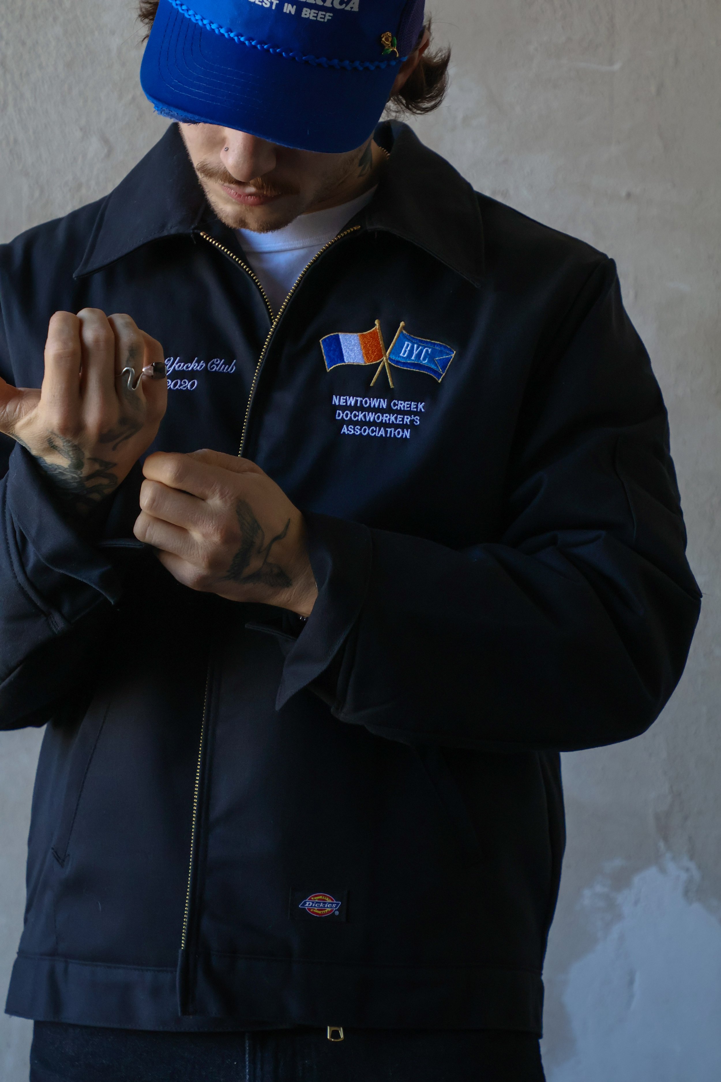 A man wearing a blue cap and a black jacket with embroidered patches, including flags and text, adjusting his jacket's zipper.