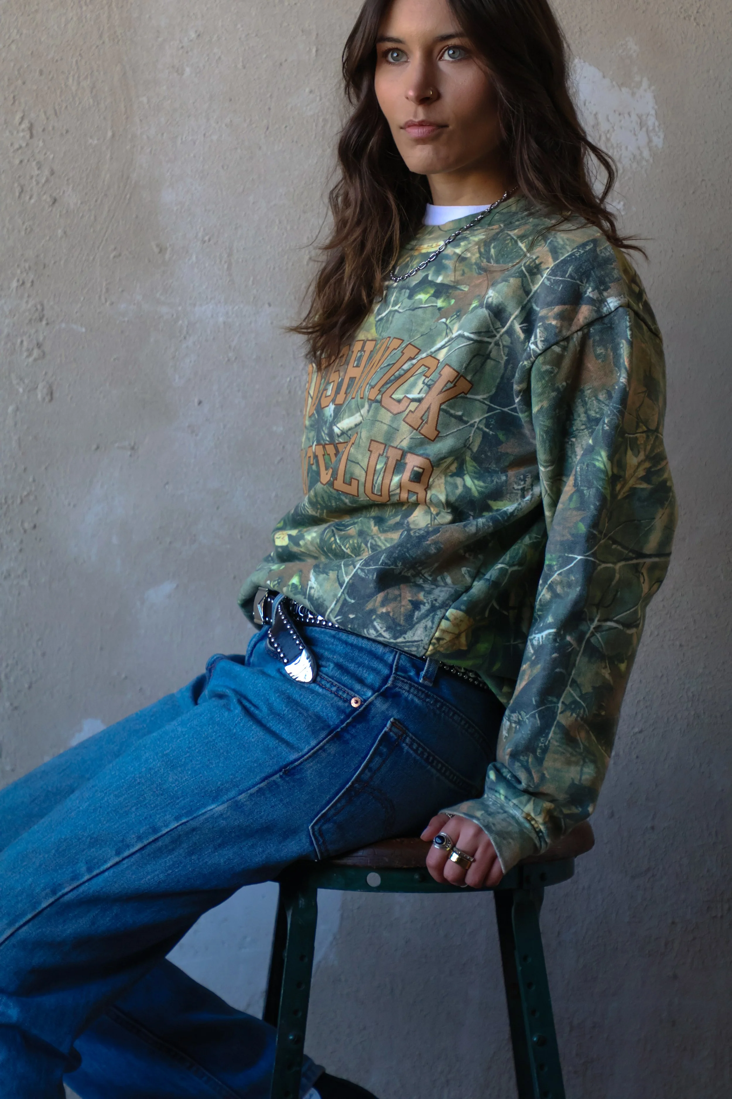 A young woman with brown wavy hair, blue eyes, and a nose ring, wearing a camouflage sweatshirt with text and blue jeans, sitting on a black chair against a textured beige wall.