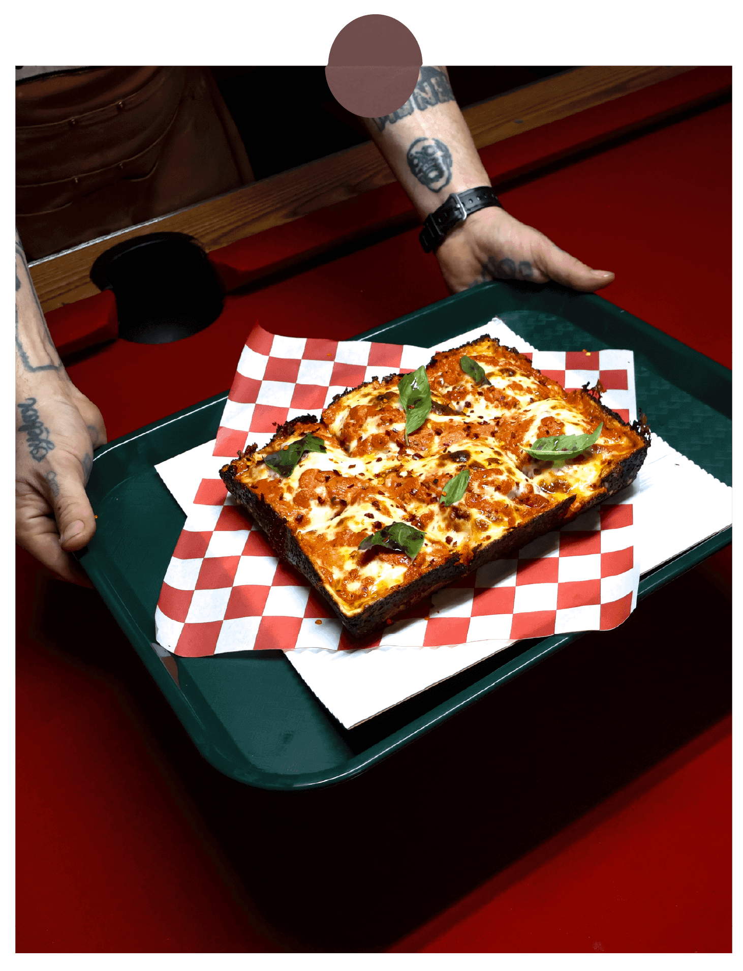A person with tattoos serving a tray holding a rectangular slice of pizza with cheese, tomato sauce, and basil leaves on red-and-white checkered paper, on a green tray.