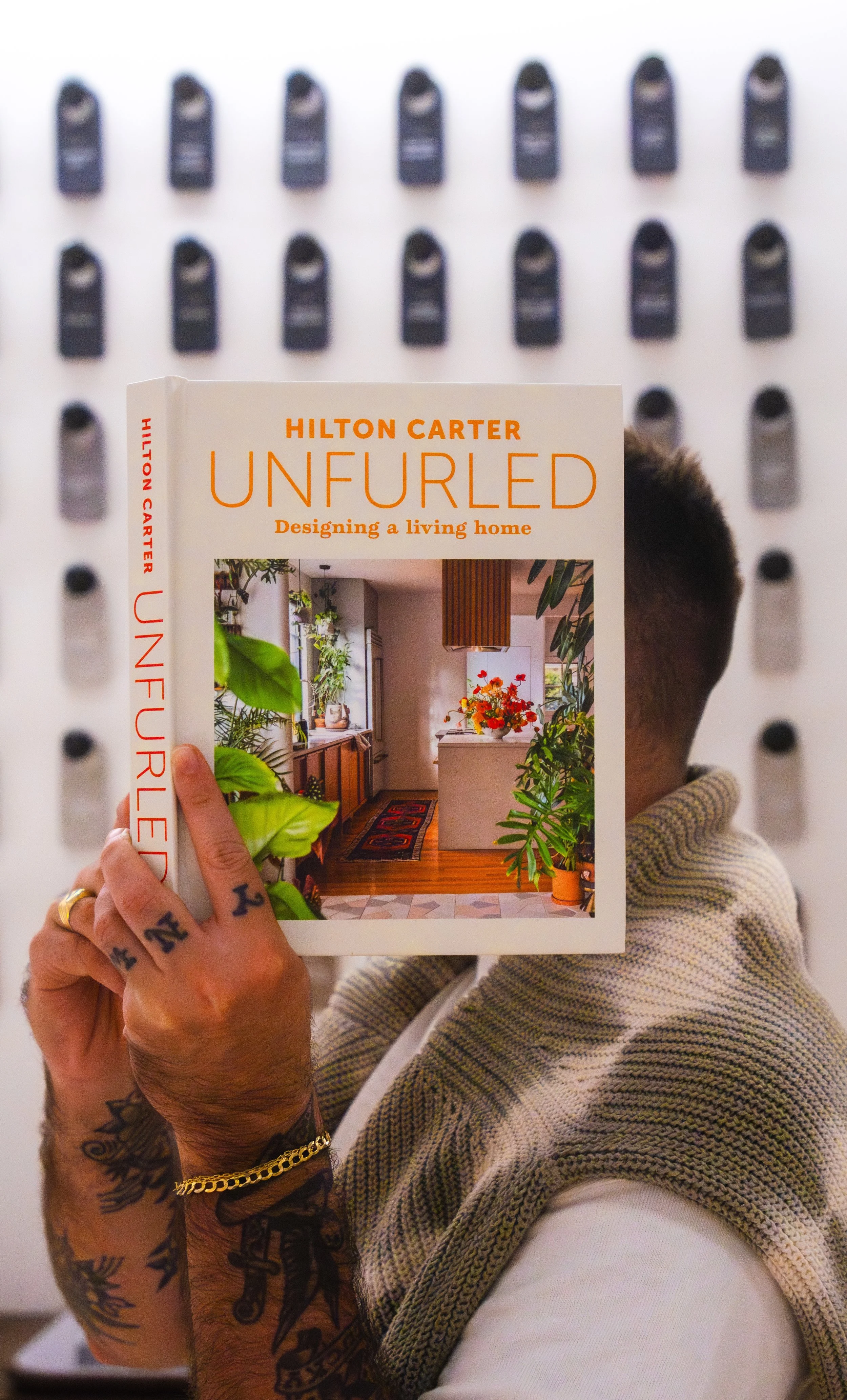 A person with tattoos on their arms holding a book titled "Unfurled" by Hilton Carter, featuring an interior design photo of a home with plants and wooden accents.