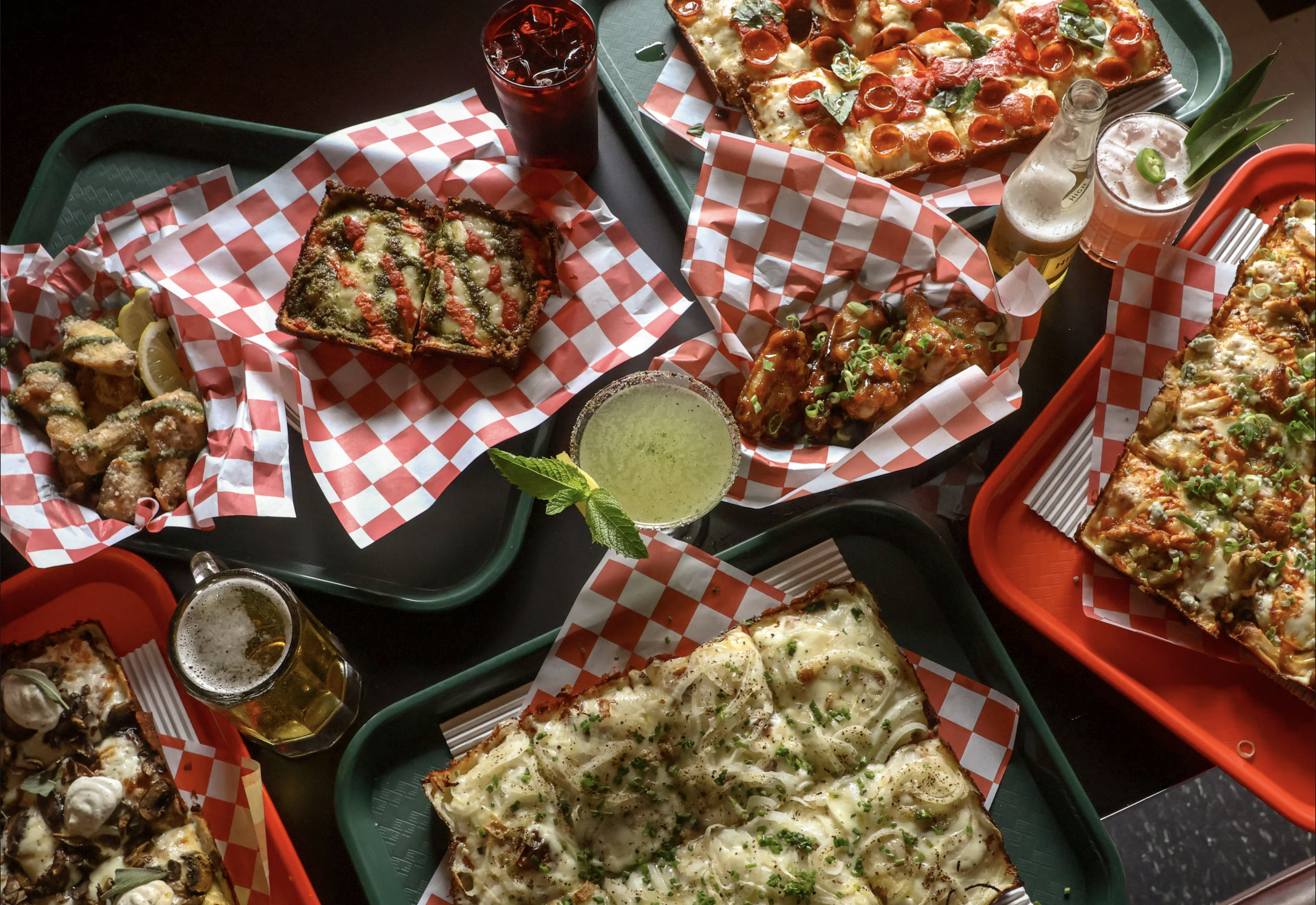 Variety of pizza slices with different toppings on red checkered paper, drinks including soda and beer, and a small bowl of green sauce, all on green trays.