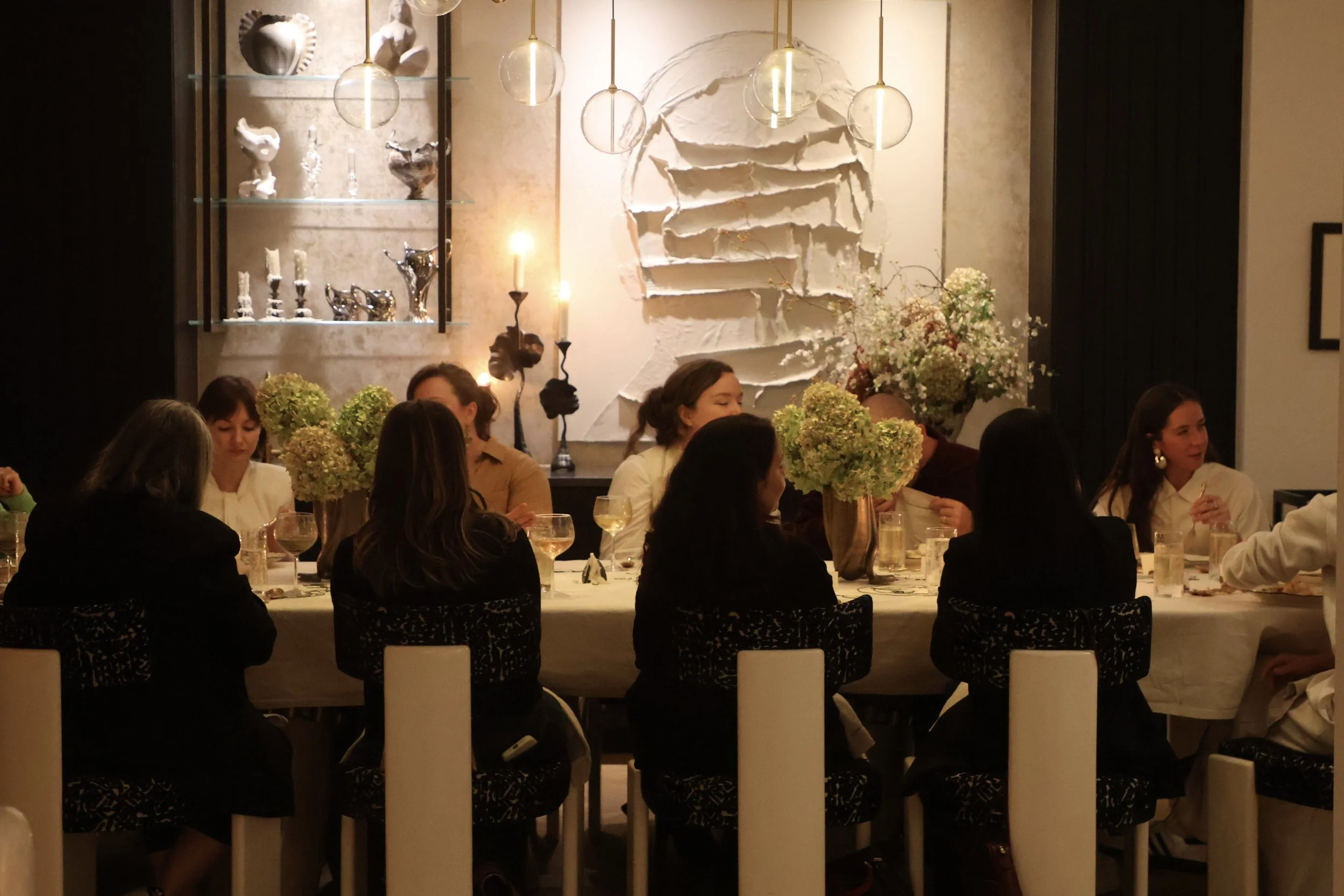 People sitting at a dining table for a dinner event in a stylish restaurant with modern decor, including abstract wall art, glass pendant lights, and floral centerpieces.