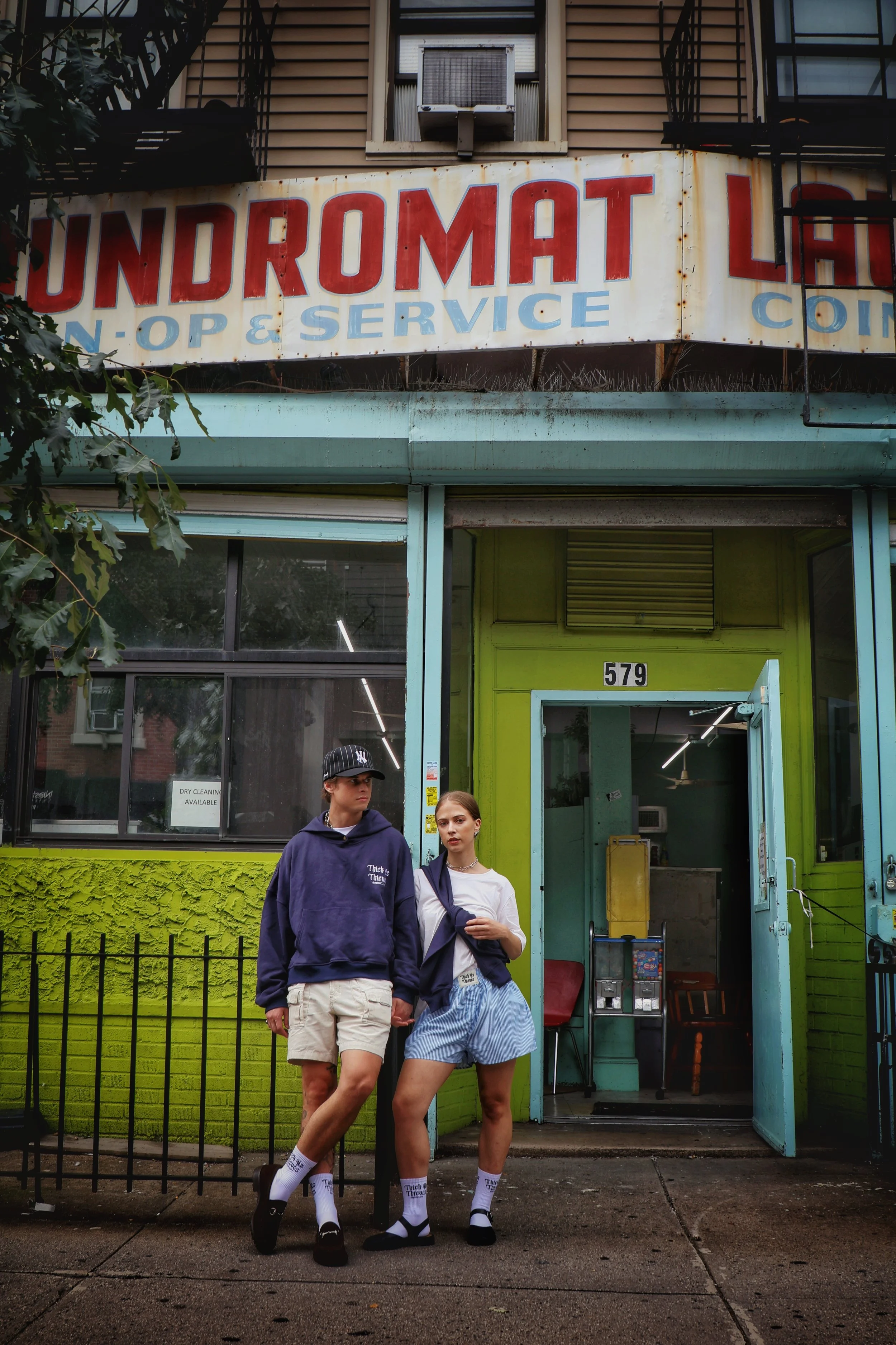 Two young people standing in front of a neon green building with an open door. The man is wearing a dark baseball cap, hoodie, and shorts, while the woman is wearing a white hoodie, striped skirt, and socks. They are holding hands and standing side by side, looking at the camera.