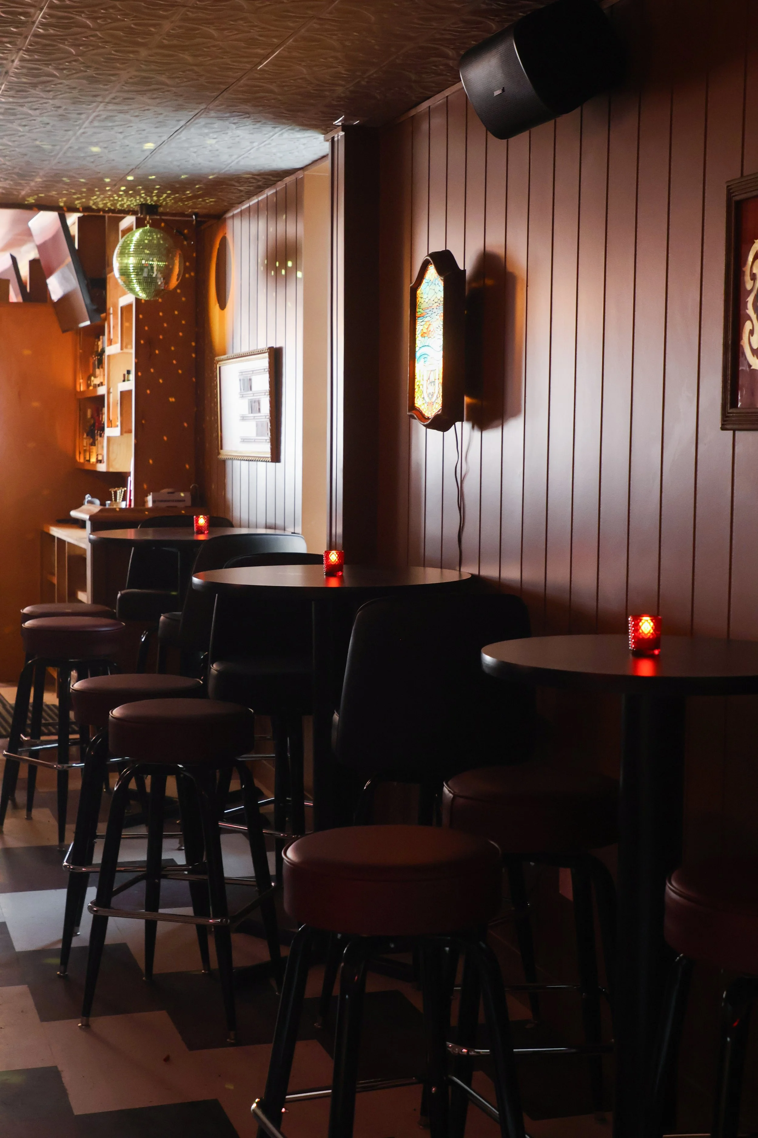 Empty bar or restaurant with black chairs, red cushioned seats, small tables with red candle holders, wooden wall panels, a stained glass decoration, and a disco ball with light reflections on the ceiling.