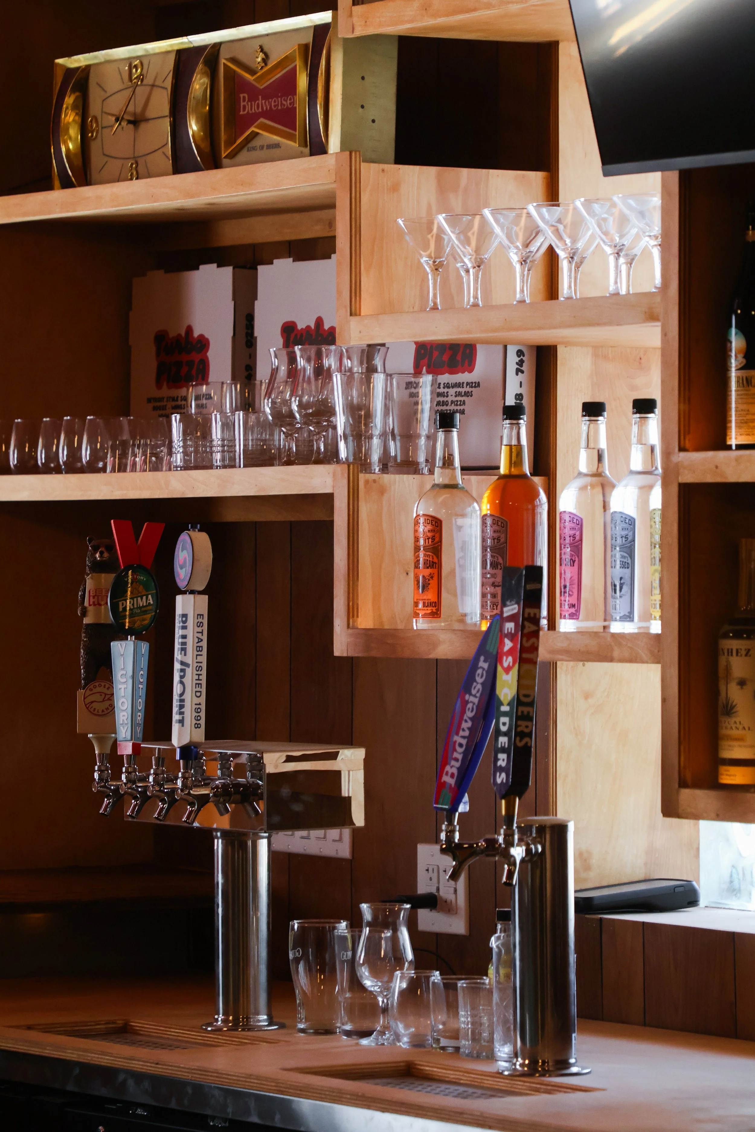 A bar with wooden shelves holding glasses, bottles, and a clock, with beer taps and glassware on the counter.