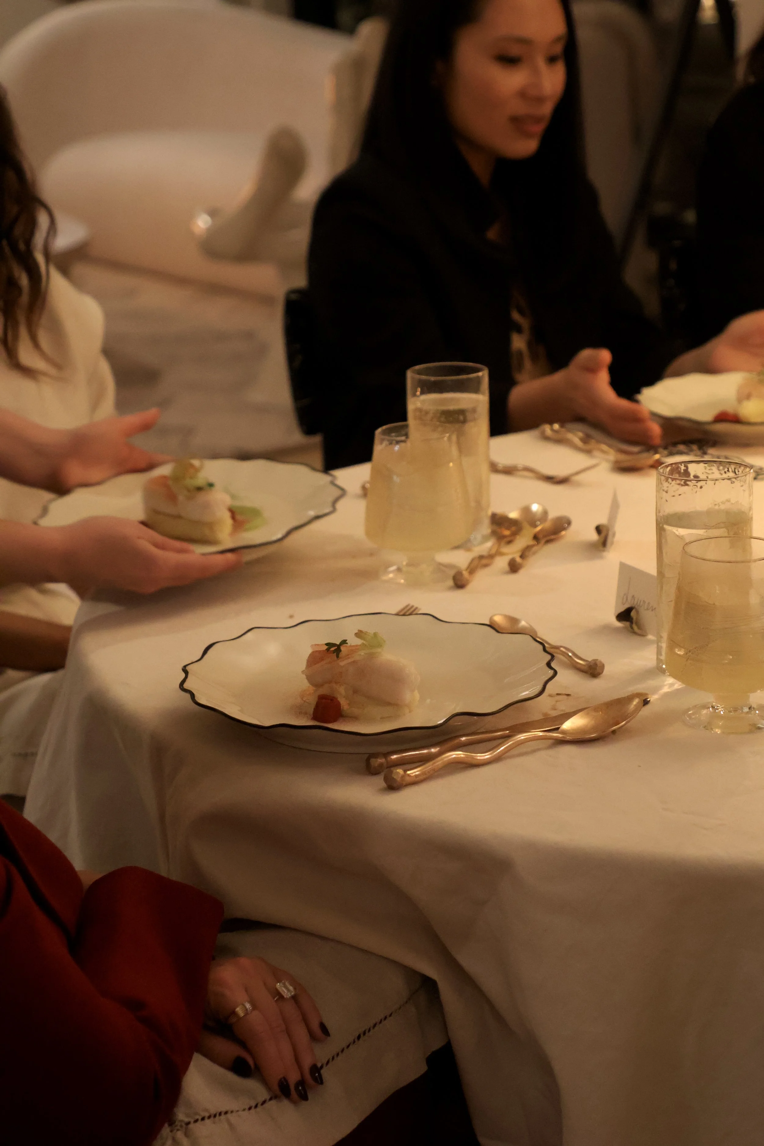 People sitting at a dinner table with plates of food, glasses of drinks, and gold-colored utensils, in a dimly lit room.