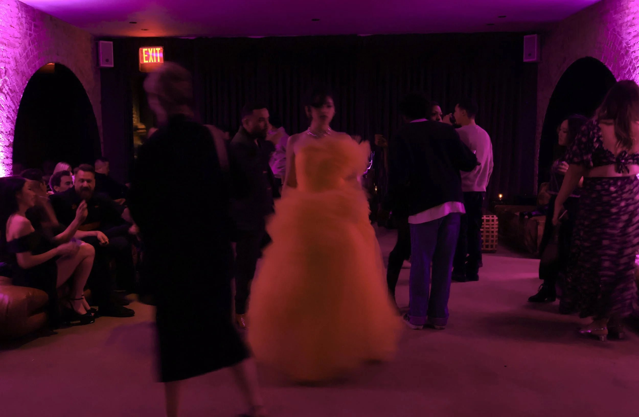 People dancing in a dimly lit room with purple lighting and exposed brick walls, some seated and some standing, with a woman in a yellow dress dancing in the center.