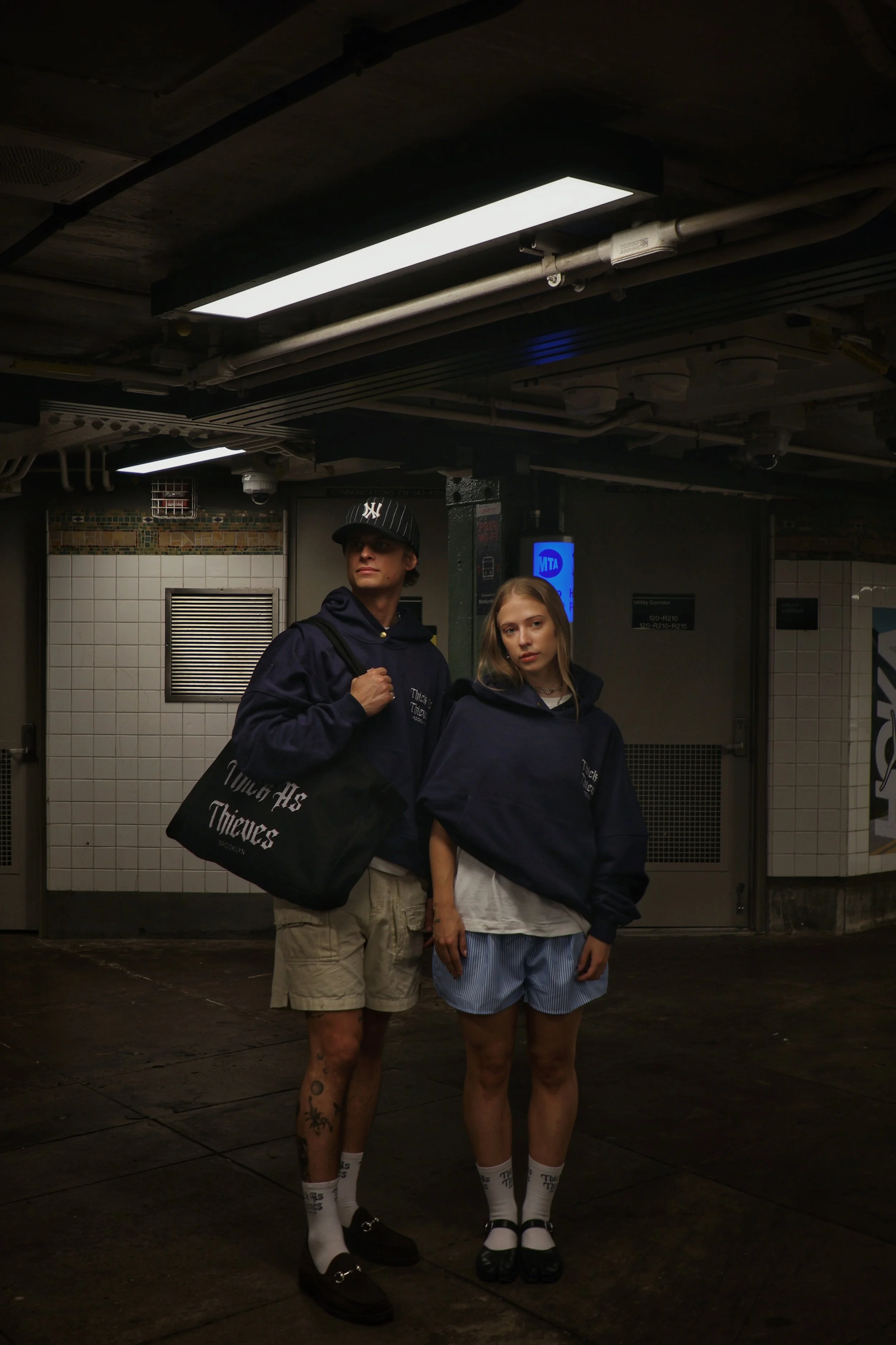 Two young individuals standing in a subway station, posing for the photo. The male on the left is wearing a black baseball cap, navy hoodie, beige shorts, and black shoes and has tattoos on his legs. The female on the right has blonde hair, and is dressed in a navy hoodie, a white shirt, and blue shorts, wearing white socks and black shoes.