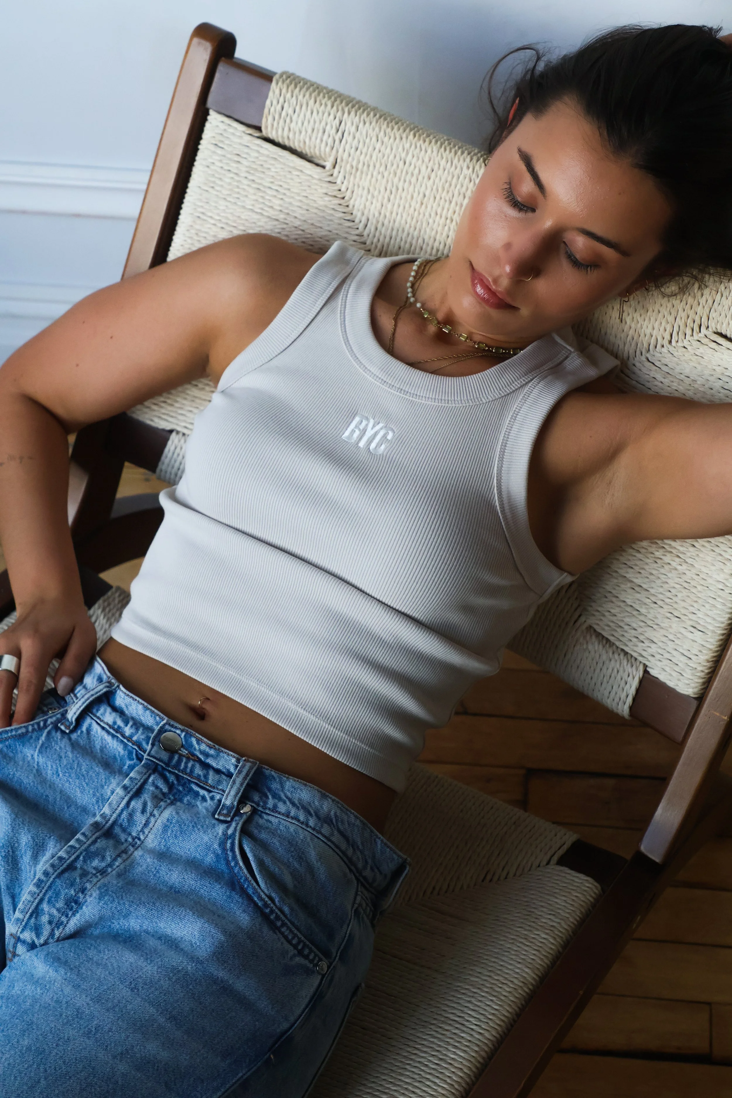 Young woman with dark hair lying on a chair, eyes closed, wearing a white tank top with 'NYC' letters and denim jeans, accessorized with layered necklaces.