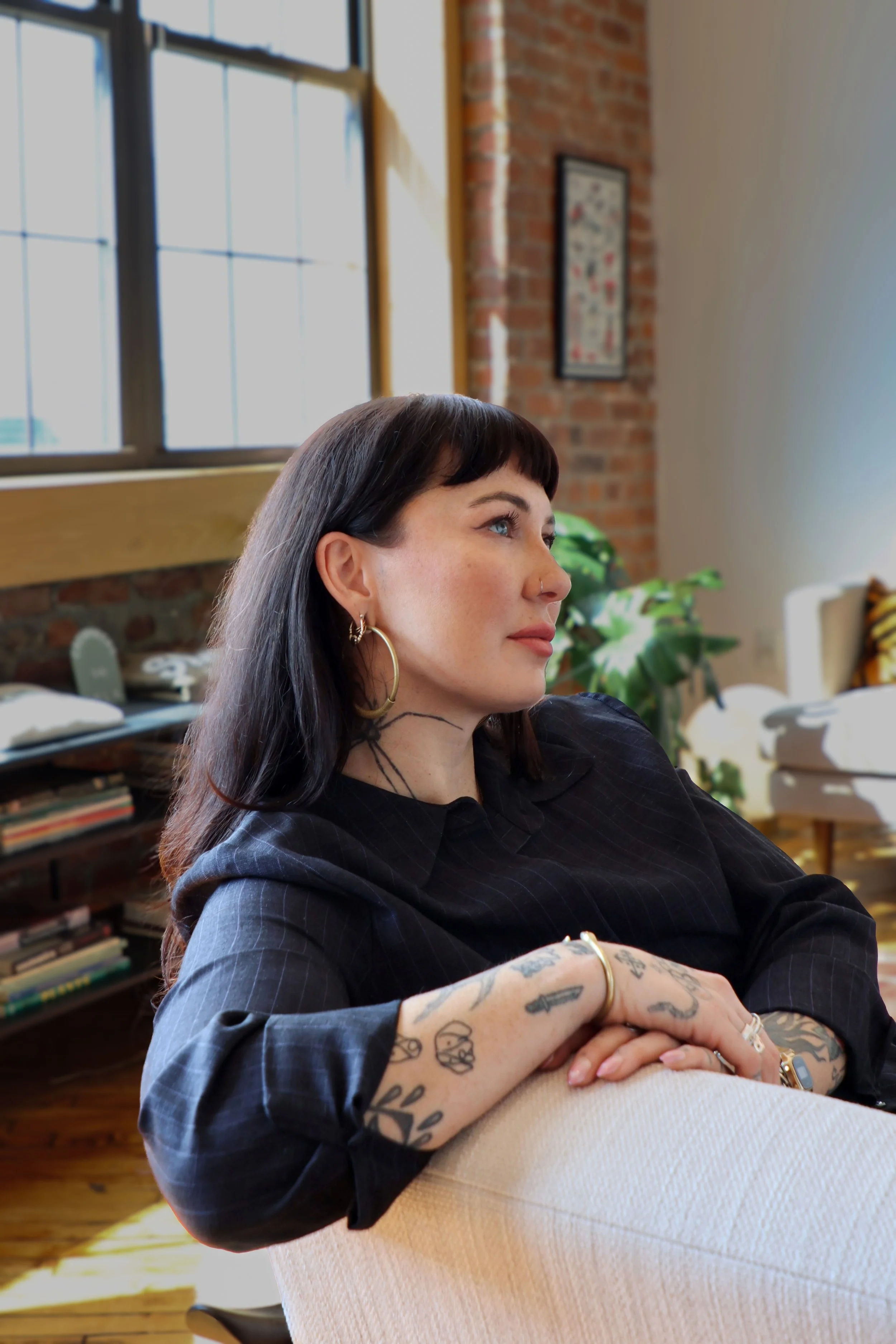 A young woman with dark hair and tattoos on her arms, wearing hoop earrings and a black shirt, is sitting indoors near large windows with a brick wall behind her and a bookshelf in the background.