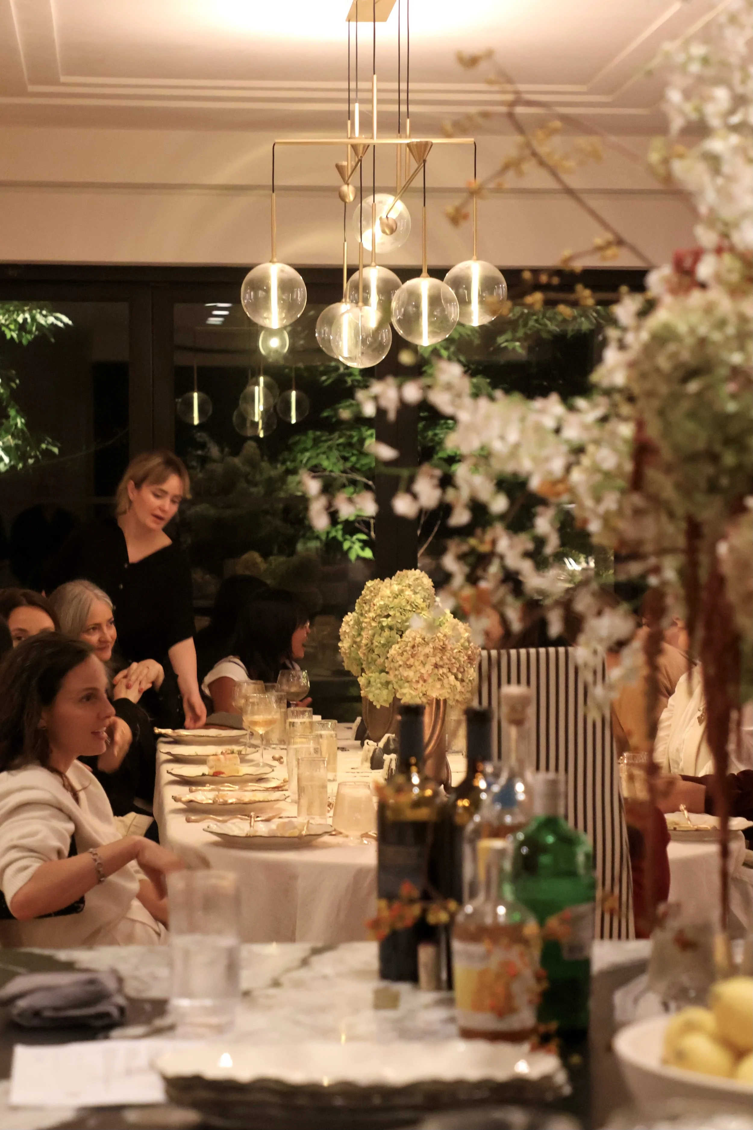 A group of people are gathered at a dining table decorated with floral centerpieces, with a modern chandelier hanging from the ceiling.