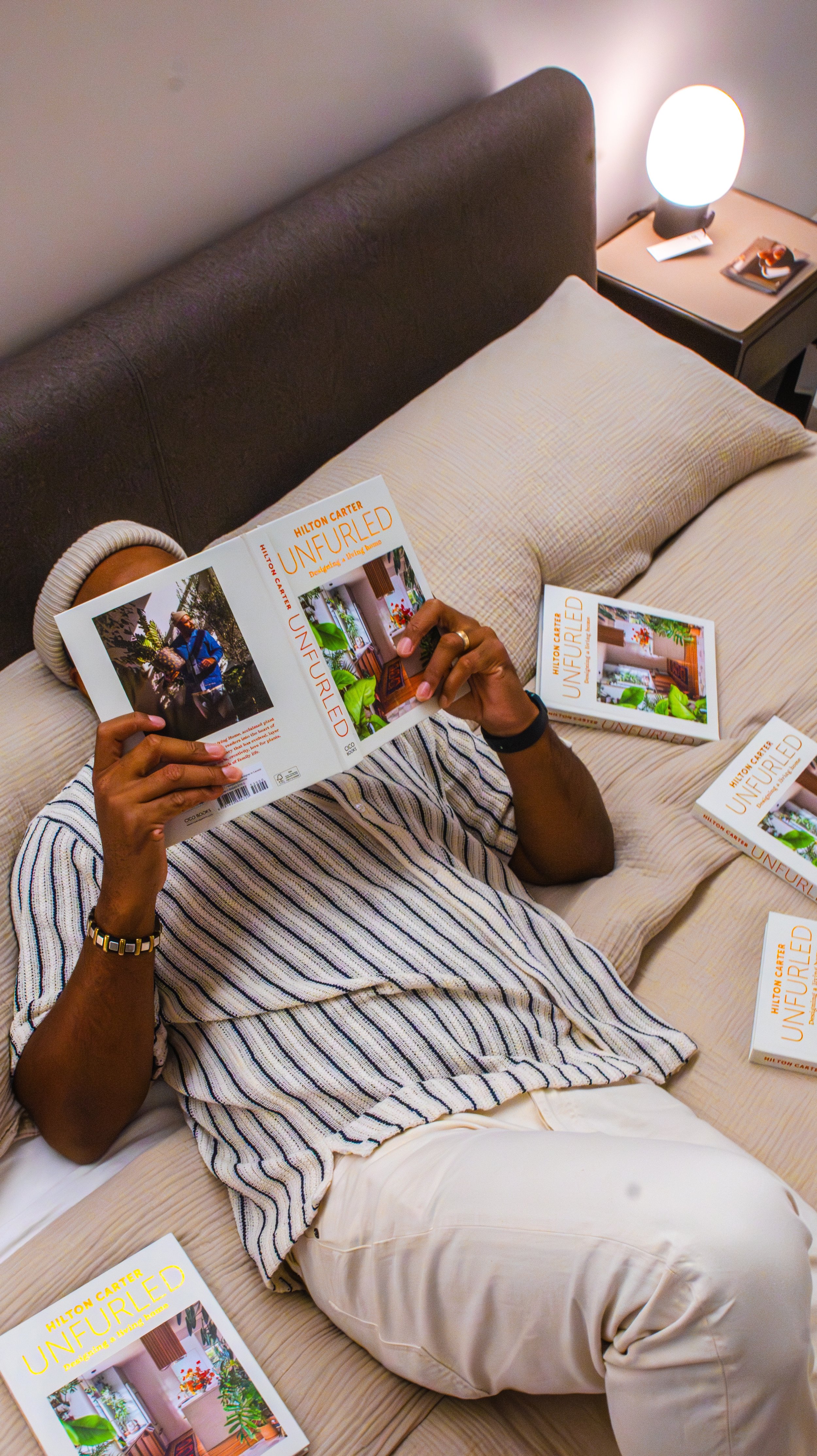 Person lying on bed reading a book titled "Unfurled" with several other copies of the book scattered around, bedside table with a lamp and small photographs, person wearing a striped shirt and light-colored pants.