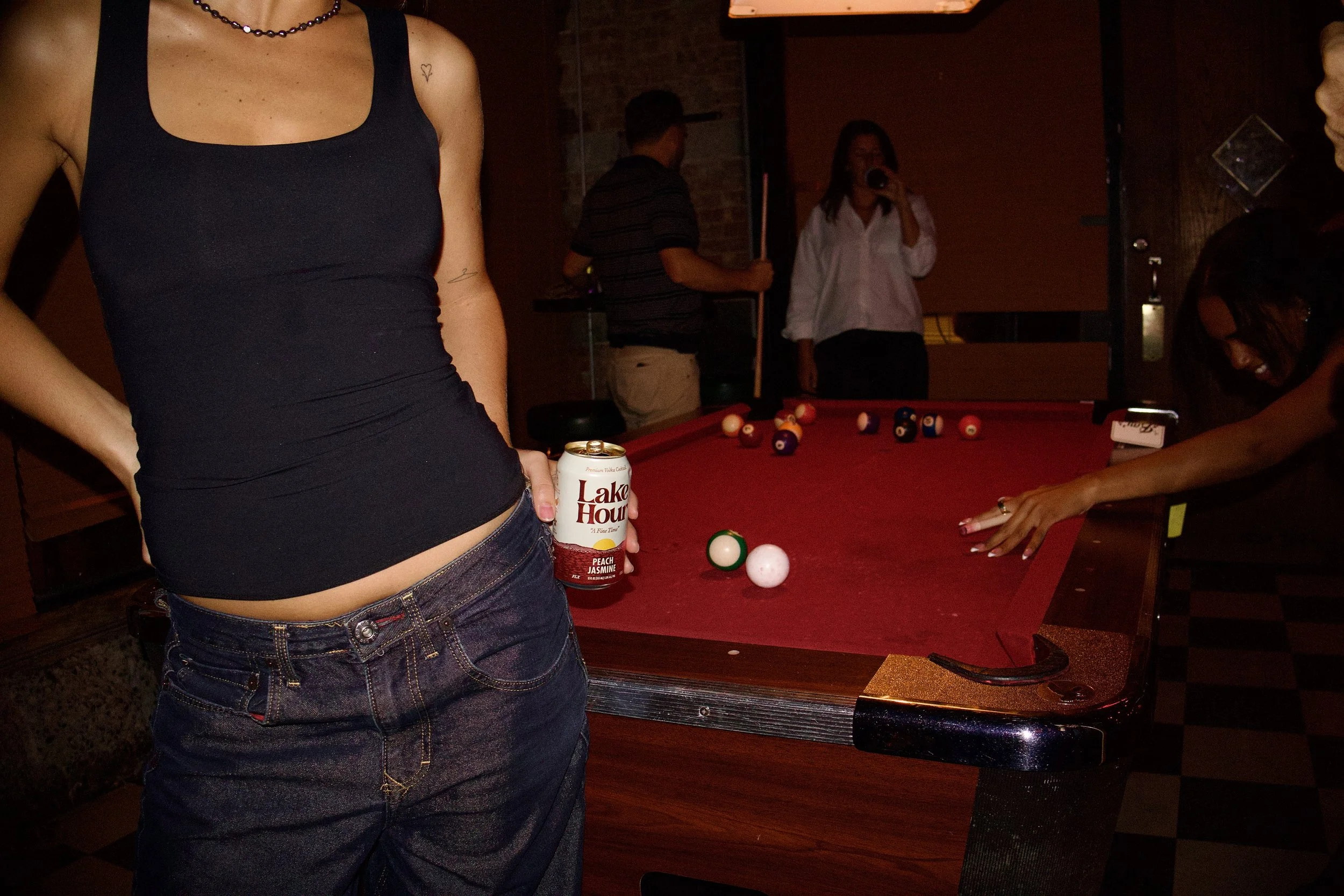 Person holding a can of Lake House Peach Jasmine flavored beverage standing near a pool table with billiard balls, while other people are in the background.