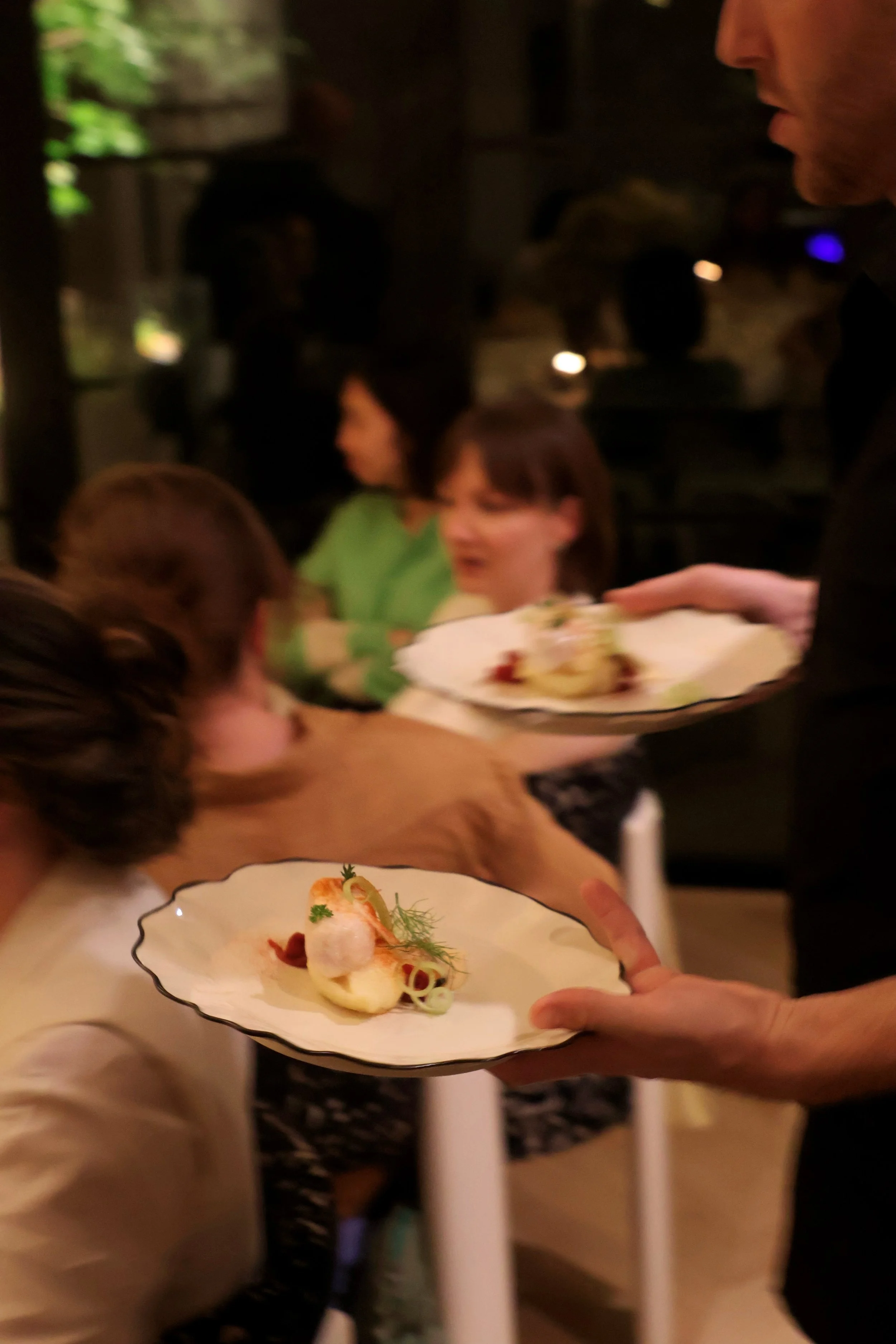 A person serving a plate of gourmet food at a restaurant event, with a focus on the elegantly plated dish featuring seafood and garnishes.