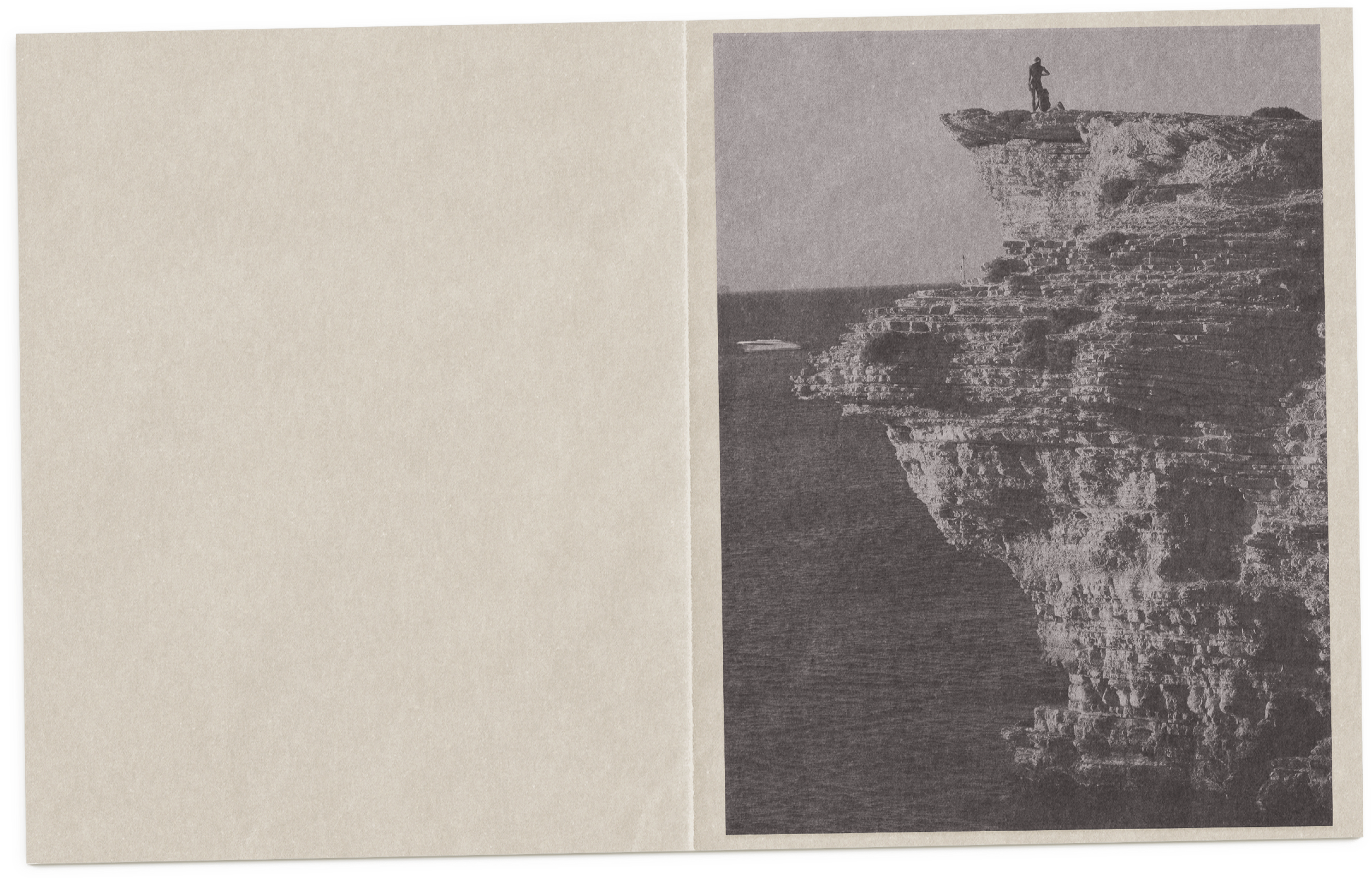 A black-and-white photograph of a rocky cliff with a person standing at the top, overlooking the water below.
