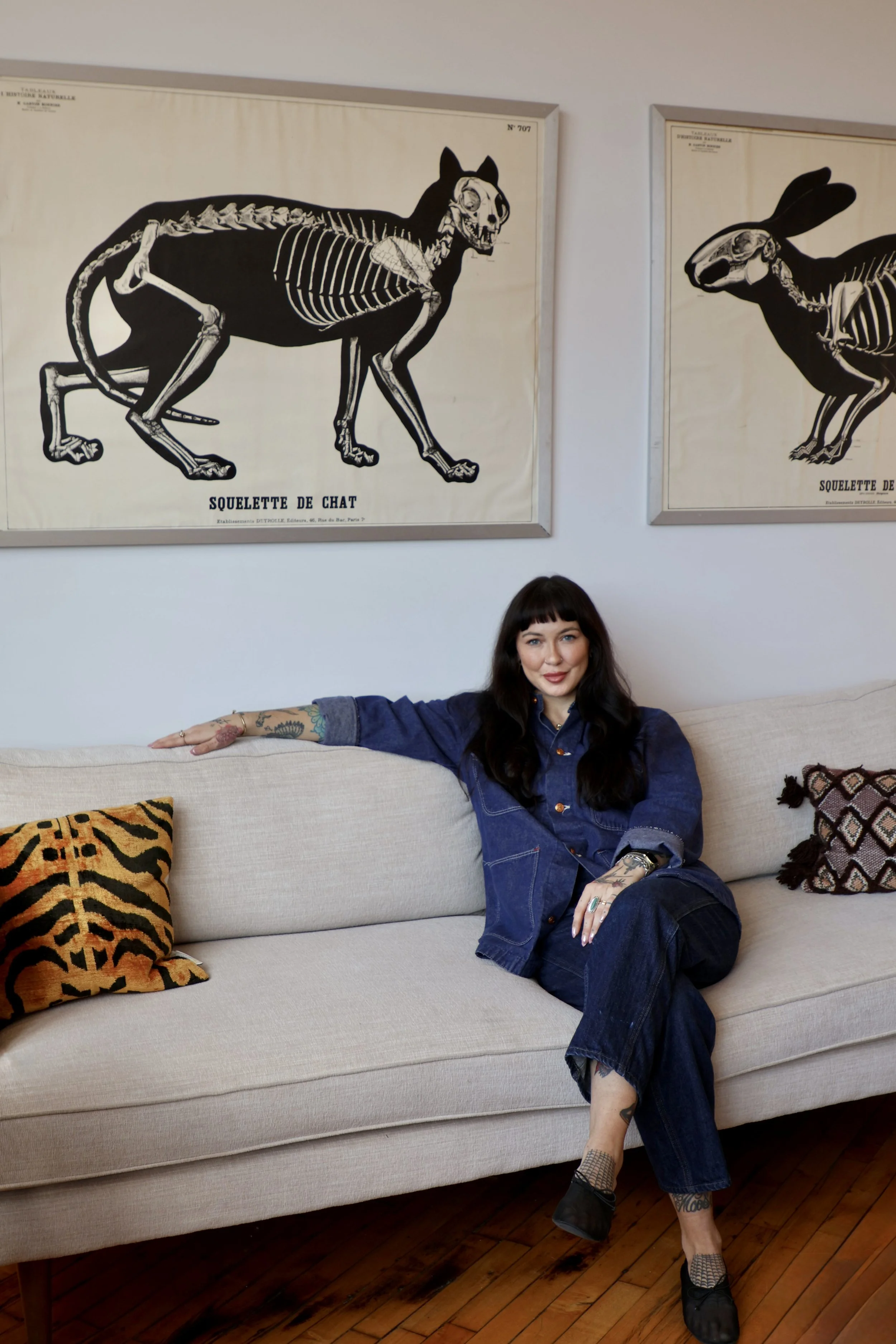 A woman with dark hair and tattoos sitting on a beige sofa, wearing a denim jacket and jeans, with a tiger print pillow and artwork of animal skeletons on the wall behind her.