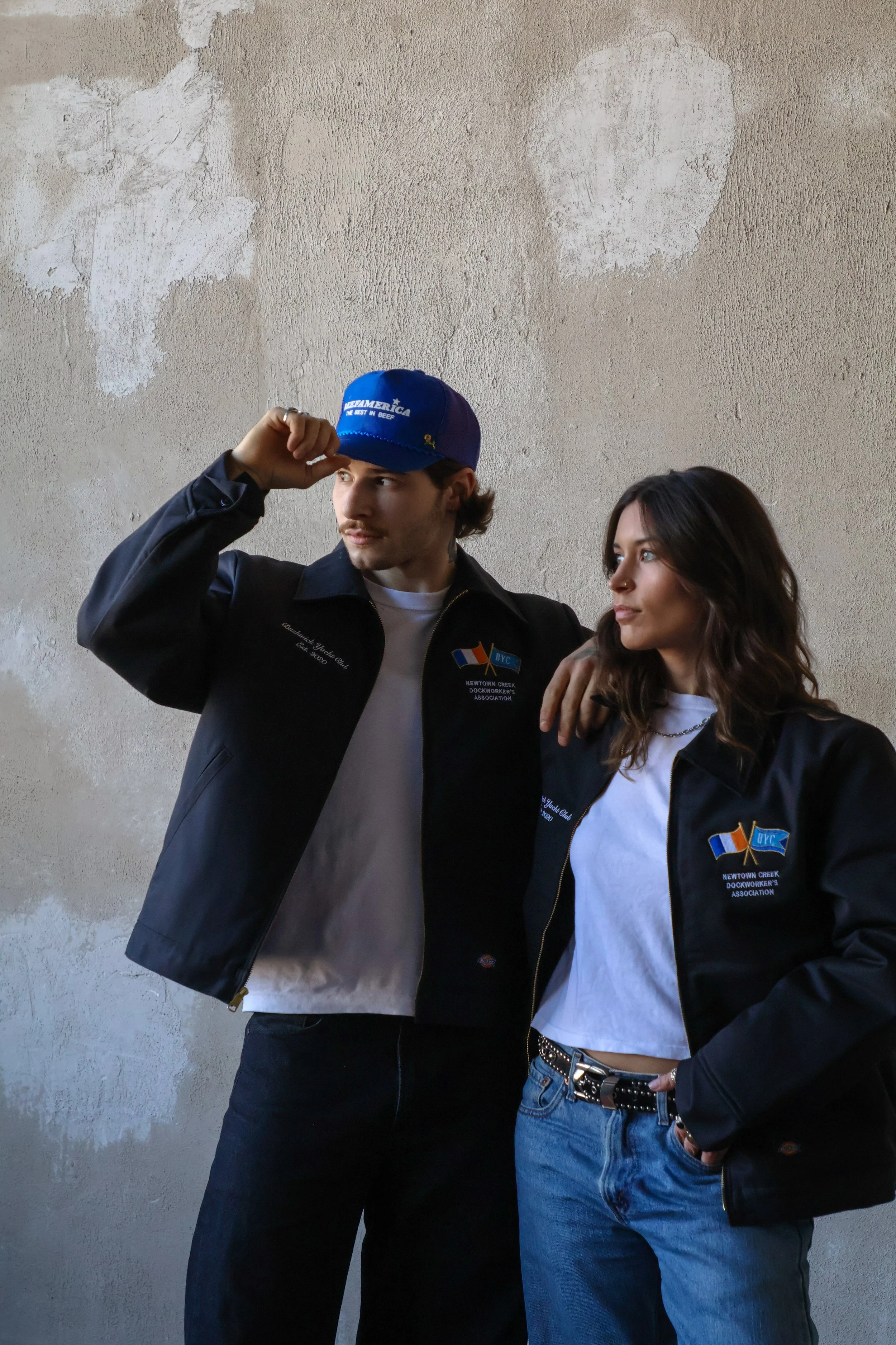 Two young adults standing against a textured wall; the male is adjusting a blue cap and wearing a black jacket with patches, while the female has her arm around him and wears a black jacket, white T-shirt, and blue jeans.