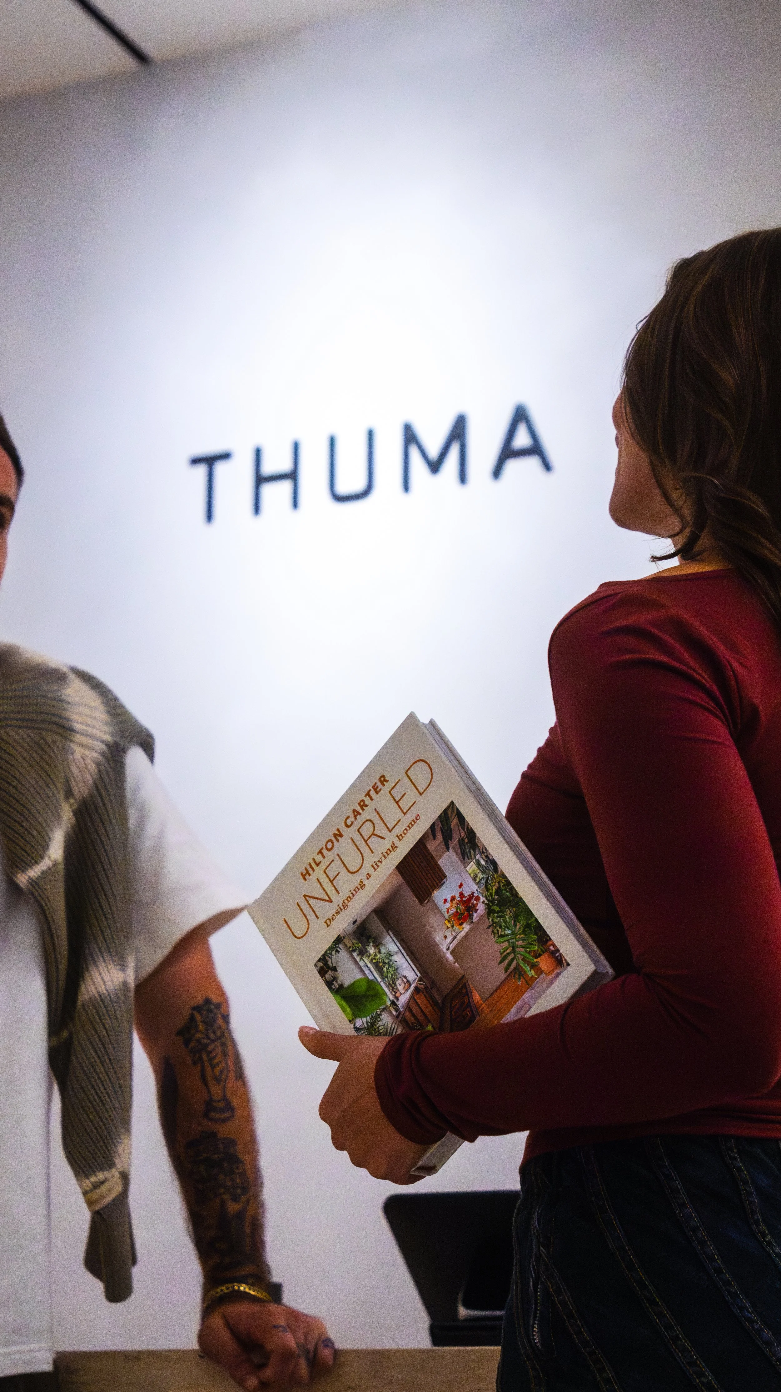 Two women indepth conversation, one holding a home decor book titled "Unfurnished" and the other looking at a sign that reads "Thuma" when seen from the back and side, in a modern interior setting.