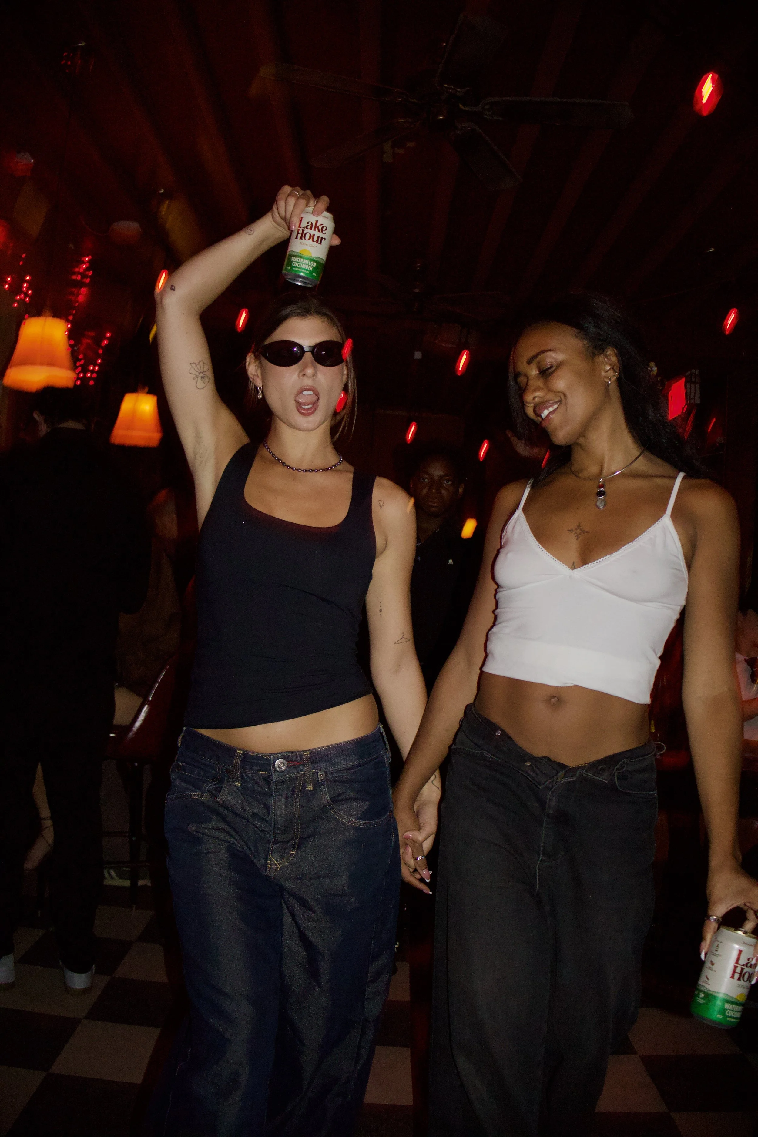 Two women holding hands in a dimly lit bar or club. One woman with long dark hair, wearing a white crop top and dark jeans, holding hands with another woman with blonde hair, wearing sunglasses, a black tank top, and jeans. The woman with sunglasses is holding a can of Lake Hous Watermelon Cucumber drink above her head and making a playful expression.