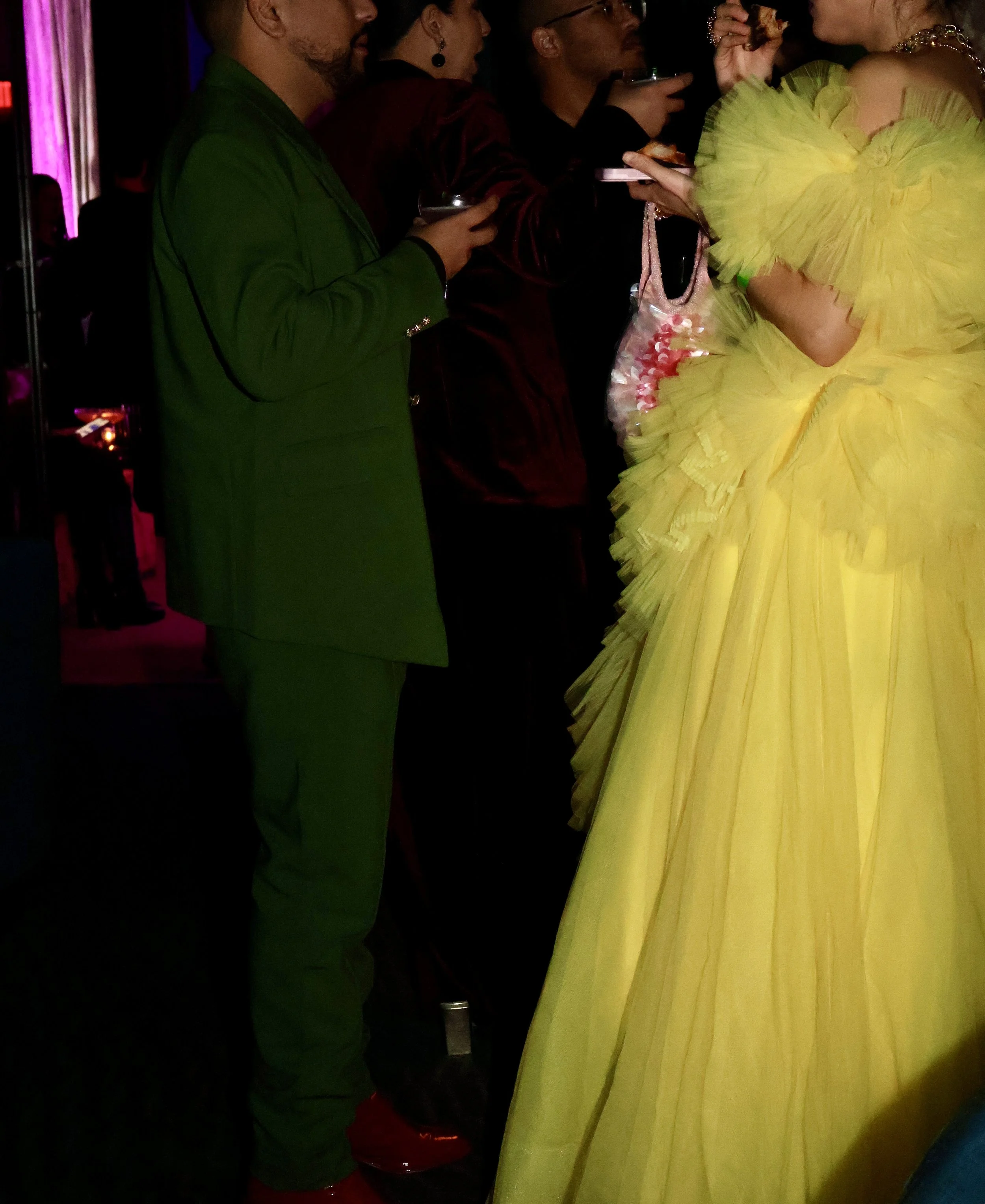 A group of people at a party, dressed in colorful and fashionable clothing, standing close together and holding drinks, with a woman in a bright yellow ruffled dress in the foreground.