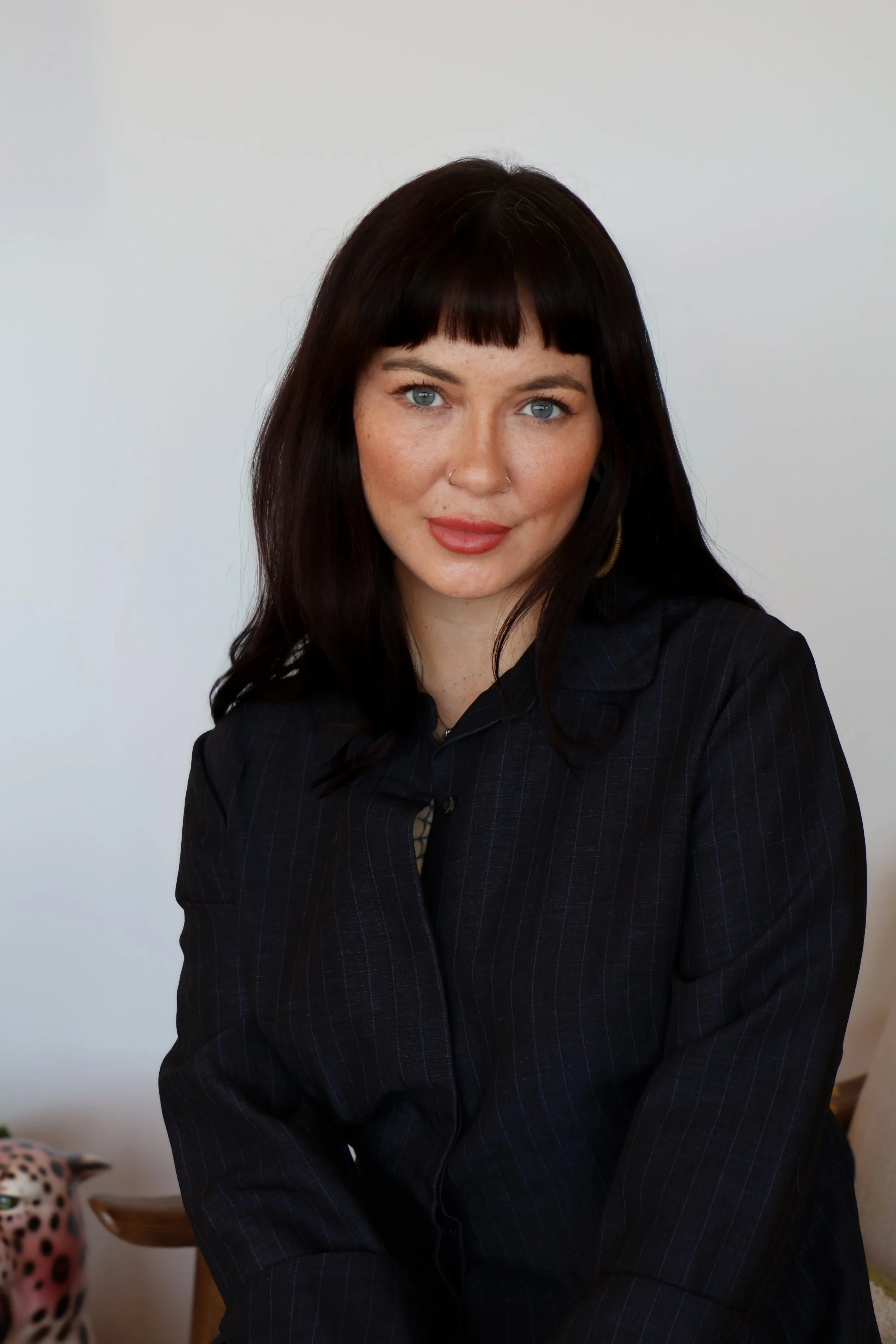A woman with black hair, blue eyes, and freckles wearing a dark pinstripe blazer sitting indoors in front of a plain white wall.