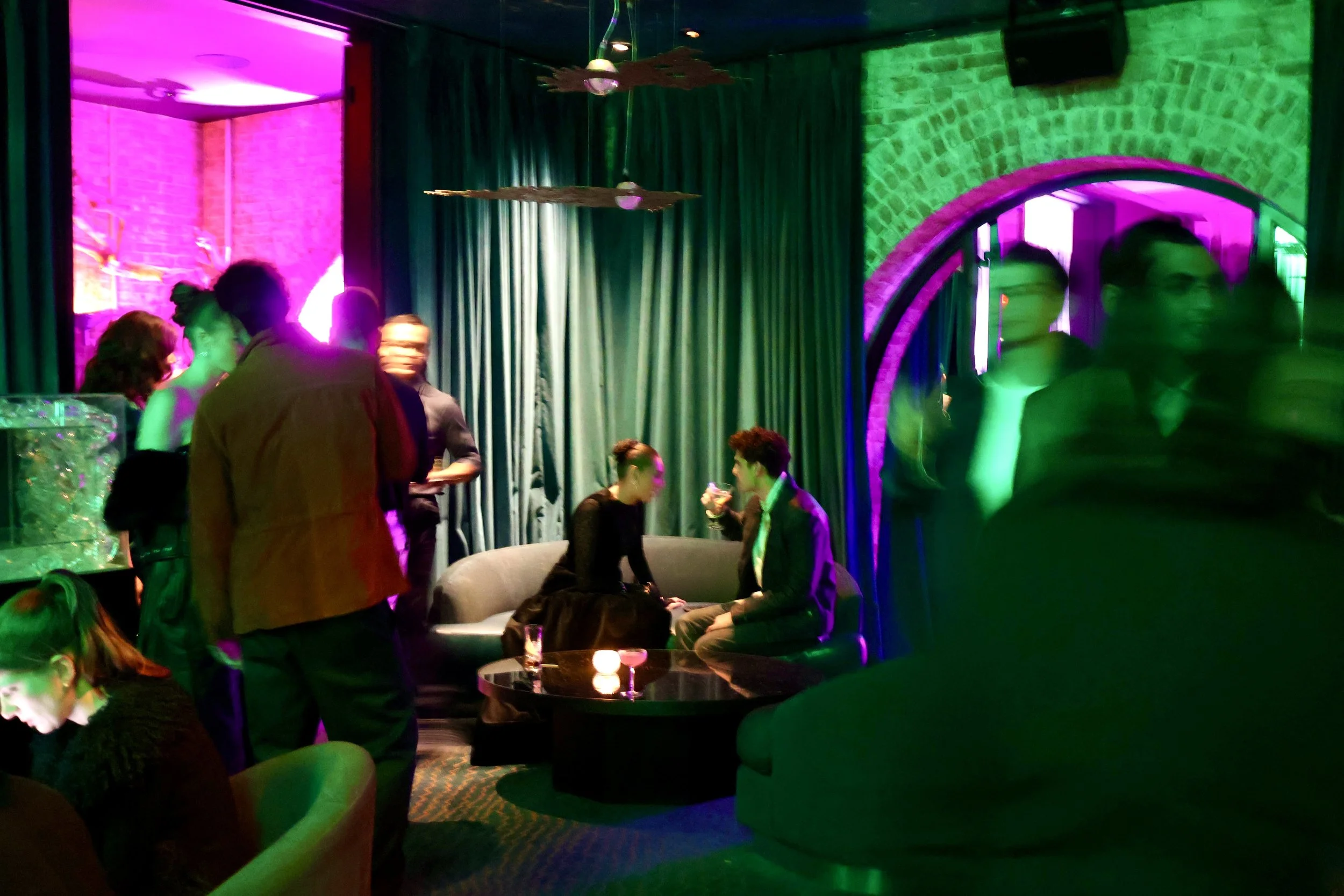 People socializing in a dimly lit lounge with colorful neon lighting, green curtains, and modern decor.