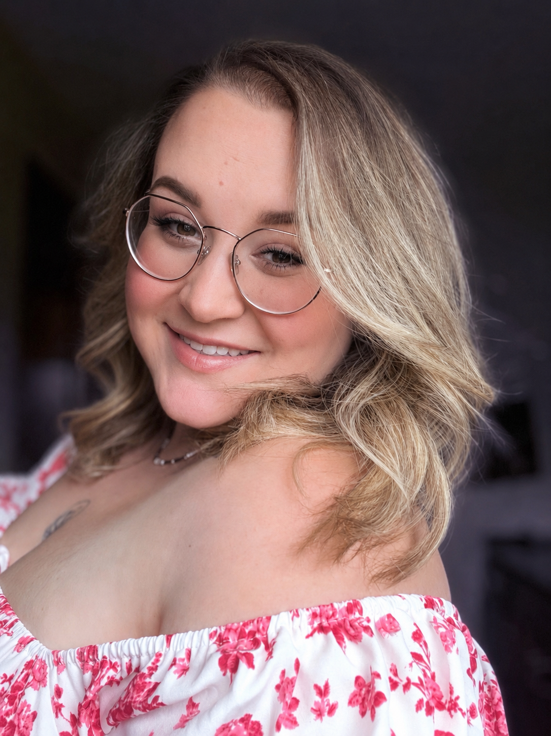 A young woman with blonde wavy hair, wearing glasses, and a floral off-shoulder top, smiling with a blurred dark background.